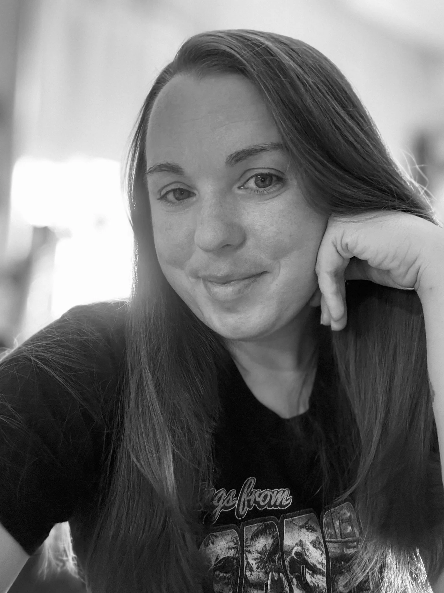 A woman with long hair and light skin resting her chin on her hand, wearing a black t-shirt with a graphic design, smiling softly in a black and white photo.