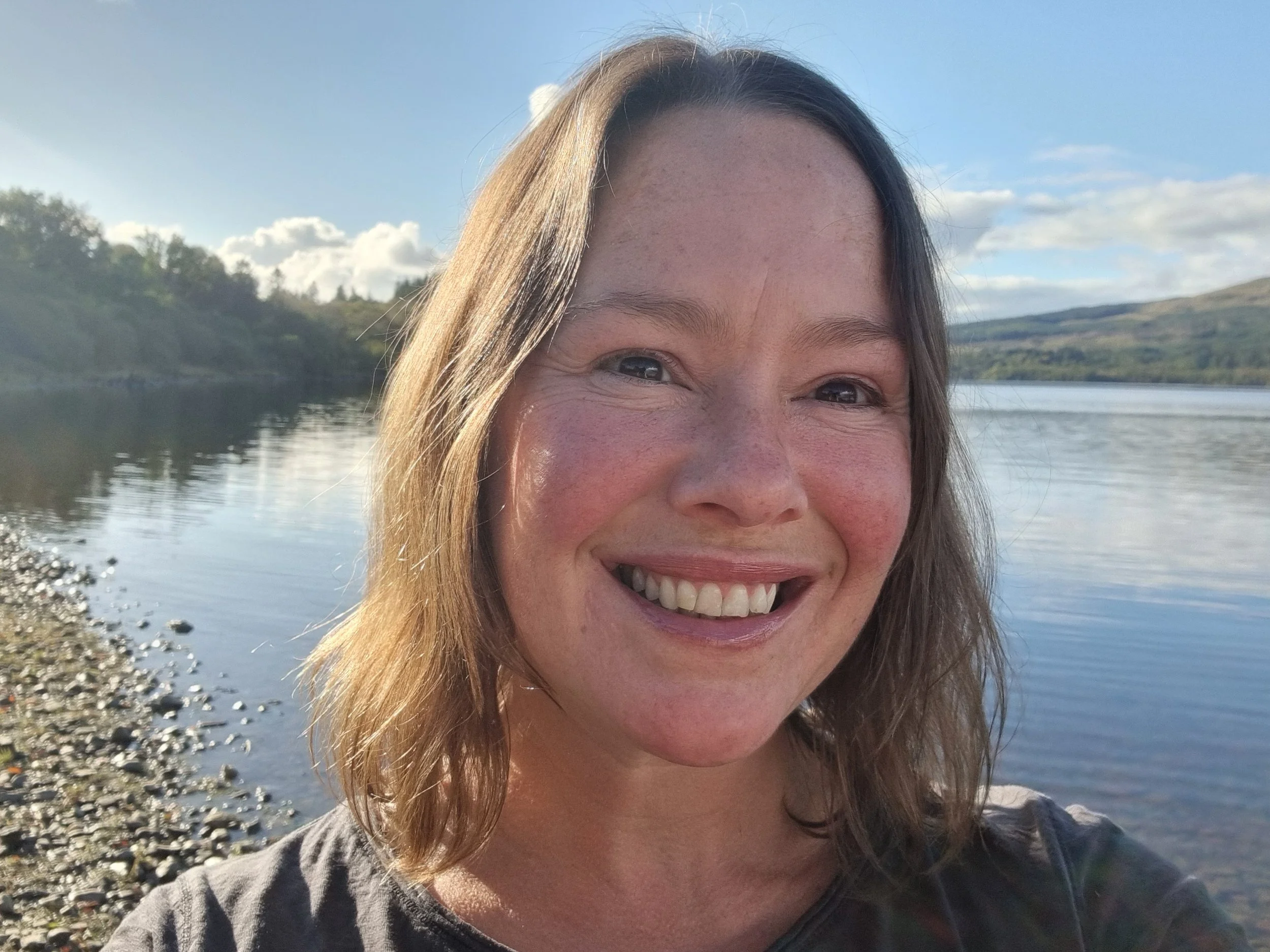 A smiling woman with long hair outdoors near a body of water, with hills and a partly cloudy sky in the background.