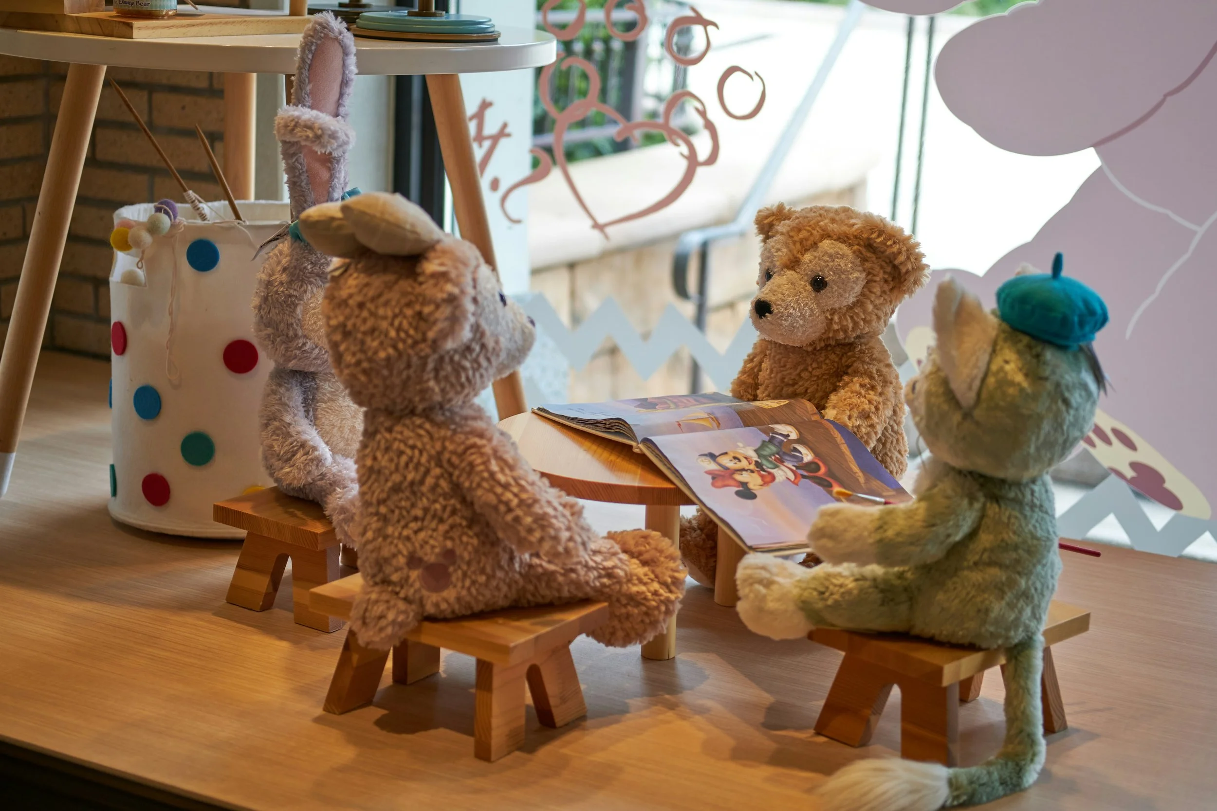 Four stuffed animals, including a bear with a yellow hat, a bear with a blue hat, and another brown bear, are sitting around a small wooden table reading a comic book. The scene is set indoors, with a display of pink flamingo decorations and a colorful polka dot container nearby.