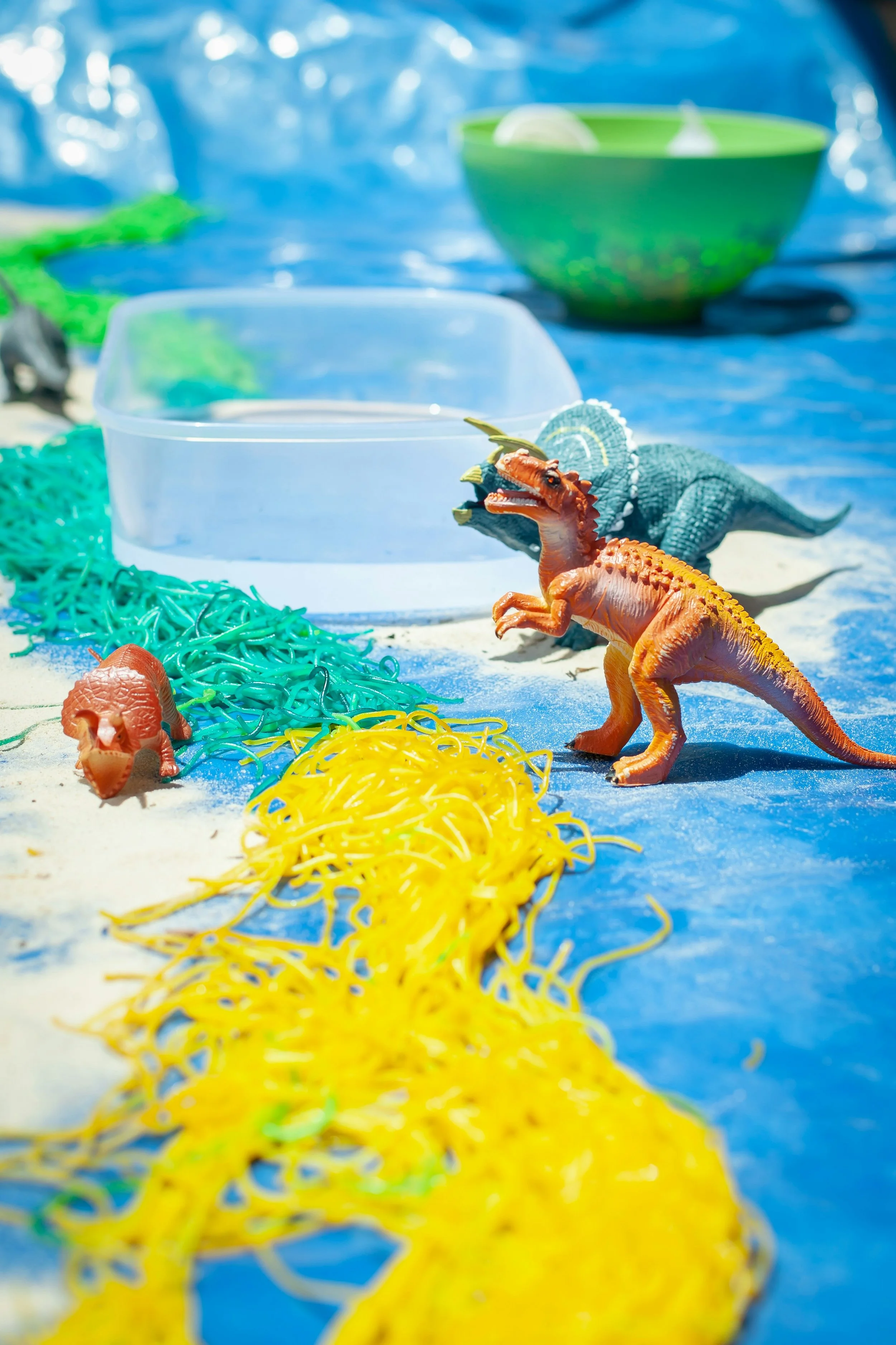 Toy dinosaurs on a blue surface with colorful rubber bands, a green bowl, and plastic containers in the background.