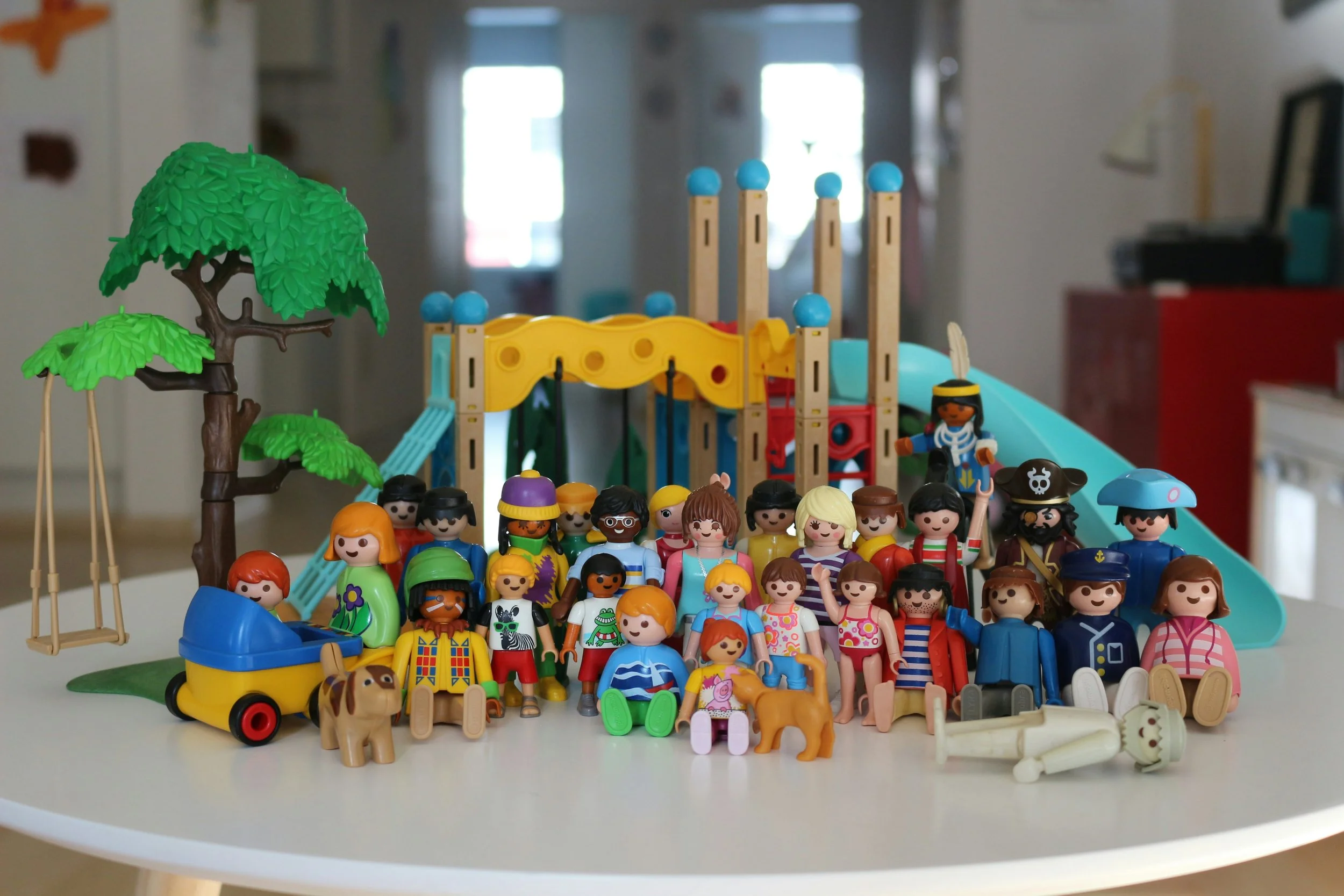 Colorful Toy figurines arranged on a white table, including trees, animals, people in various outfits, and amusement park elements like a slide and a carousel.