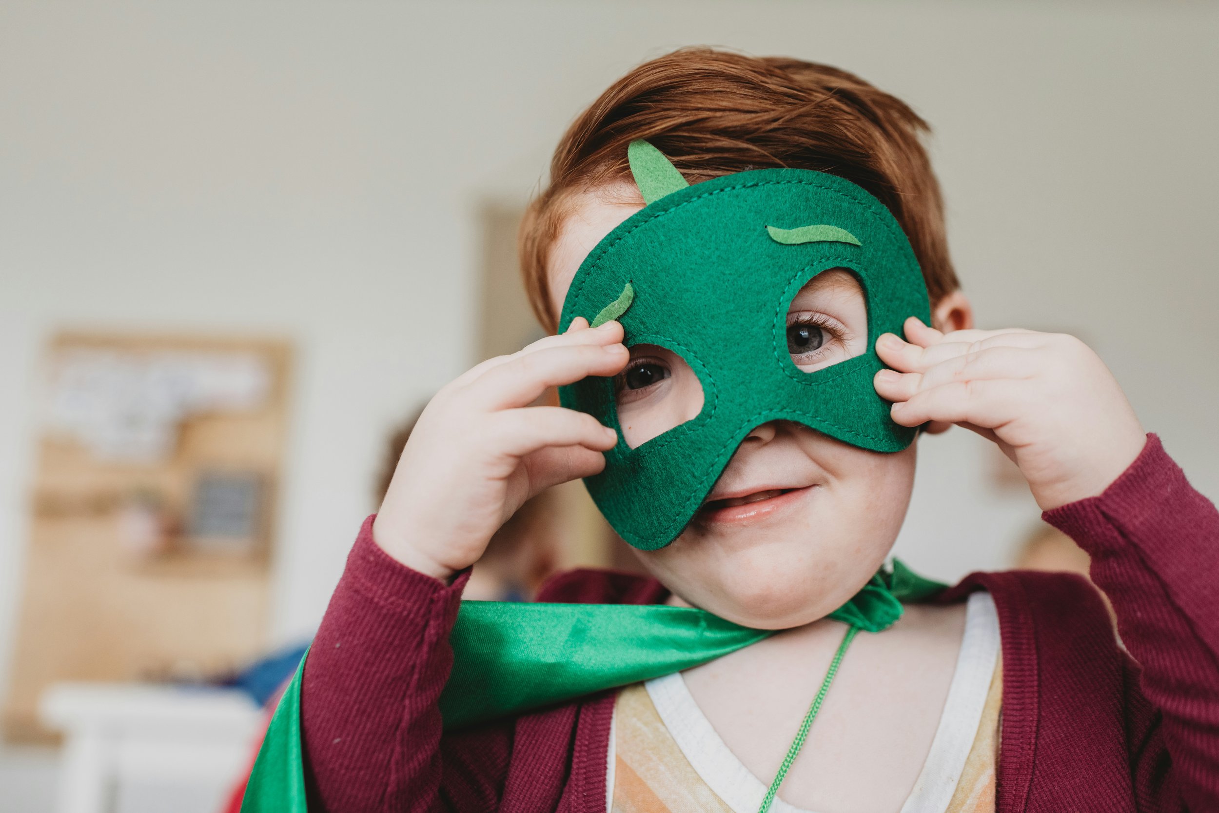 A young child dressed as a superhero, wearing a green felt mask with felt ears and leaf details, and a green cape. The child is holding the mask with both hands and looking at the camera with a smile.