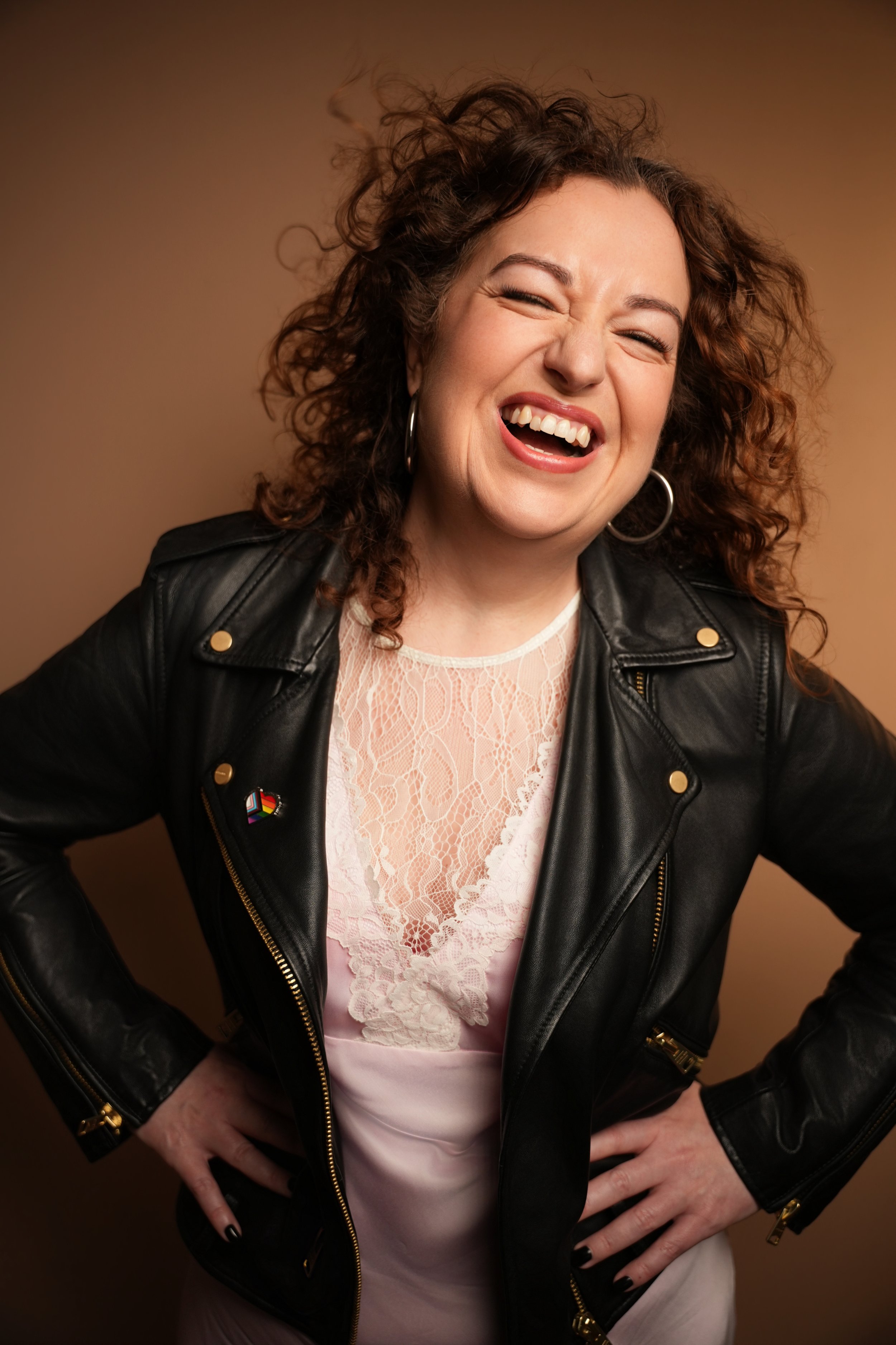 Jorden, the owner of Stay Beautiful Facial Studio smiling, wearing a black leather jacket over a pink lace dress, standing against a brown background.
