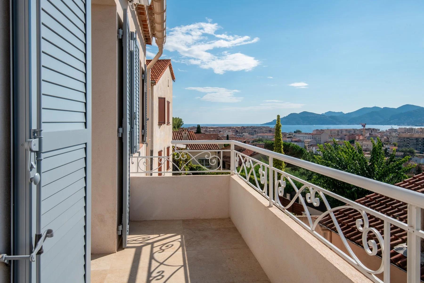 Balcon avec une vue sur la mer, montagnes en arrière-plan, et toits de maisons avec des tuiles rouges. Balustrade en fer blanc et volets bleus sur la maison.