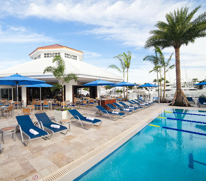 Poolside view with blue lounge chairs, umbrellas, palm trees, and a restaurant near a marina with boats.