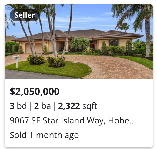 A single-story house with a brown roof, surrounded by palm trees and well-maintained landscaping, including shrubs and grass, with a curved brick driveway leading to the house.
