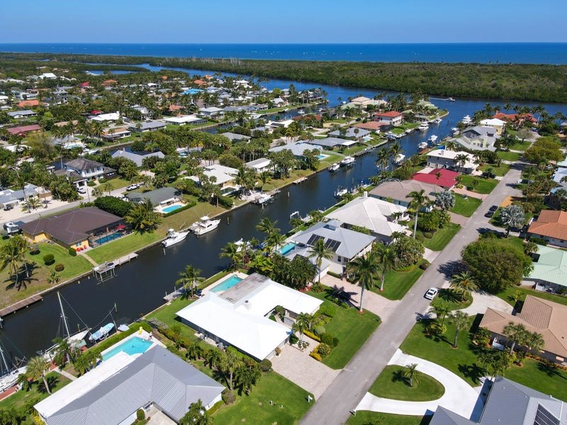 Aerial view of a coastal neighborhood with houses along a canal, boats docked, green lawns, palm trees, and the ocean in the background.