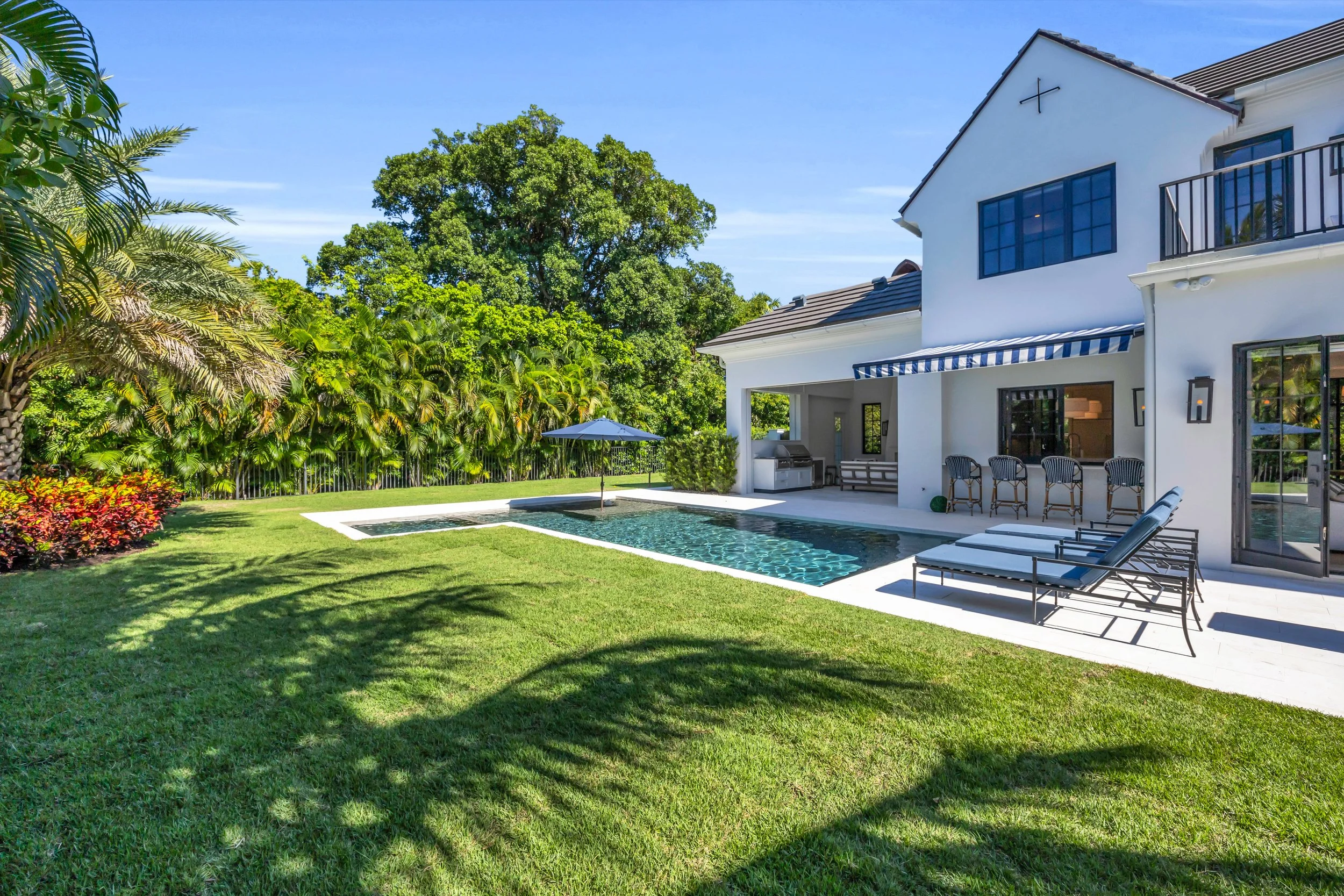 White modern house with a backyard swimming pool, surrounded by lush greenery and trees, with outdoor seating and a blue and white striped awning.