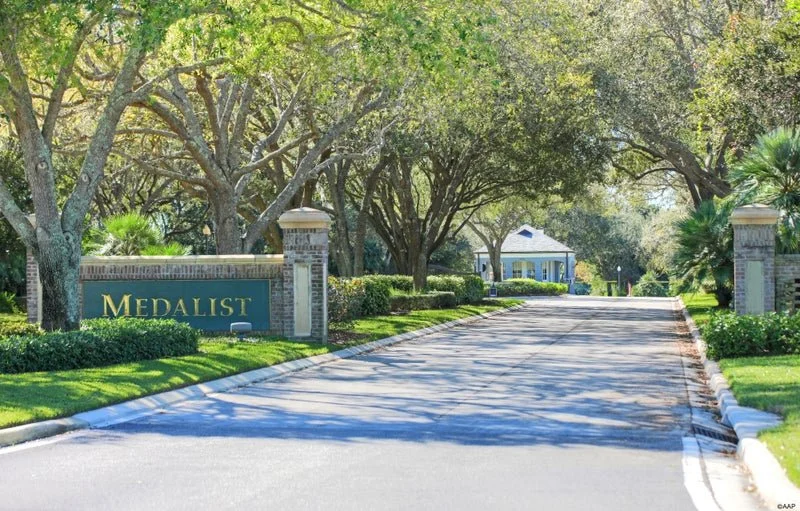 Entrance to Medalist neighborhood with a decorative sign on the left, lush green trees and landscaped bushes along the street, leading to a gated community.