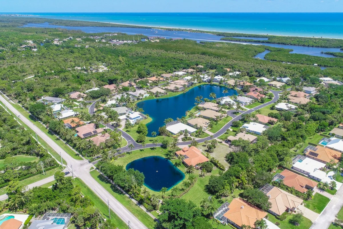 Aerial view of a residential neighborhood with houses, multiple lakes, and green trees, near the coastline with the ocean in the background.