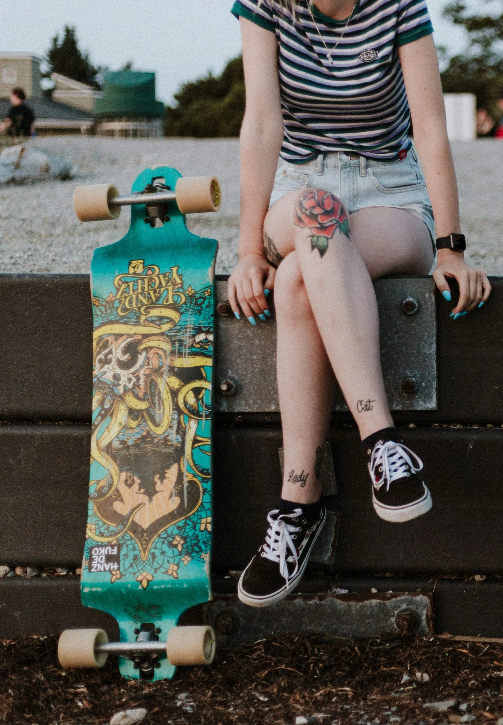 A person sitting on a skateboard at a skatepark, wearing a striped shirt, denim shorts, and holding a skateboard with a colorful design nearby. The person's tattoos read 'Cat' on one leg and 'Lady' on the other. They are wearing black and white athletic shoes and a wristwatch.
