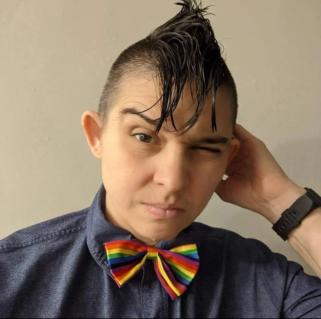 Person with short hair styled with wet-looking, spiky hair on top, wearing a denim shirt and a rainbow-striped bow tie, making a winking facial expression while touching their head with one hand.