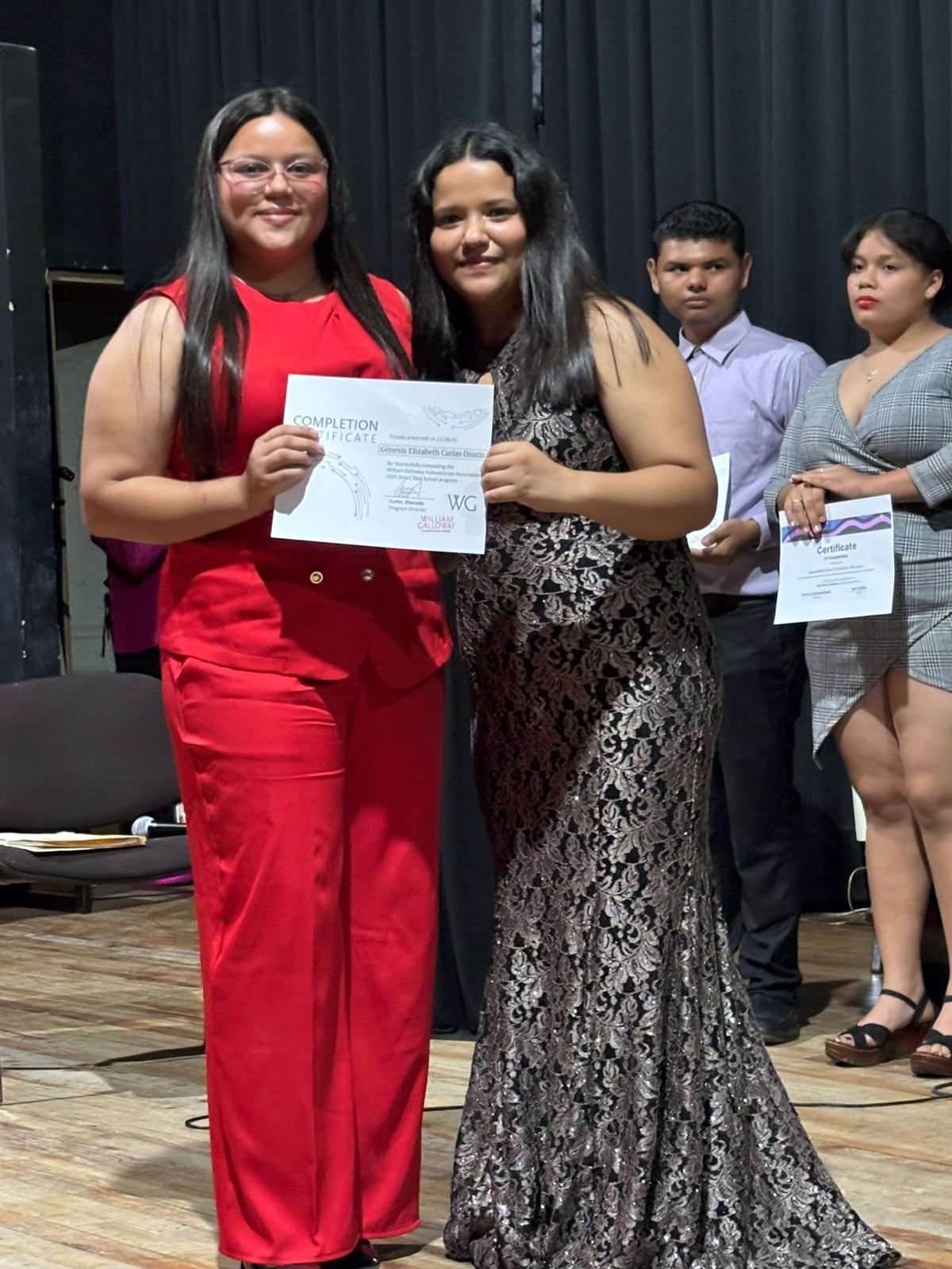 Two women standing on a stage holding a certificate and smiling, with two people in the background also holding certificates, against a black curtain backdrop.