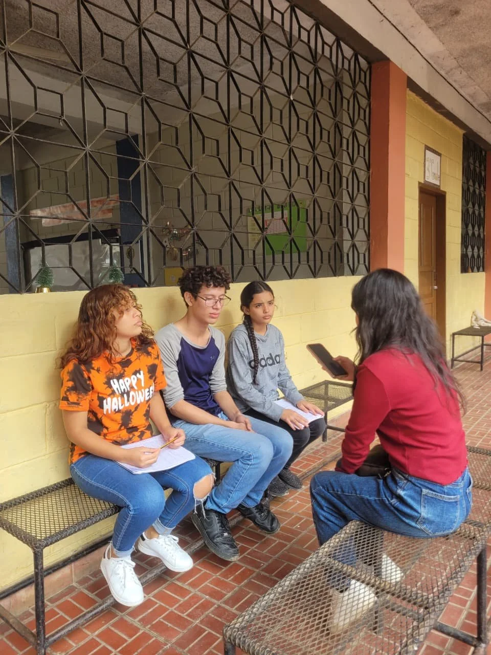 A woman is sitting on a wirebench, talking to three teenagers sitting on a yellow wall, two girls and a boy. The girl on the left is wearing an orange Halloween t-shirt, and the girl on the right is wearing a gray Adidas sweatshirt. The woman is holding a phone and appears to be interviewing or talking to the teens.