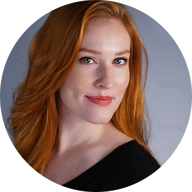 Close-up of a woman with long red hair and fair skin, smiling softly and looking at the camera.