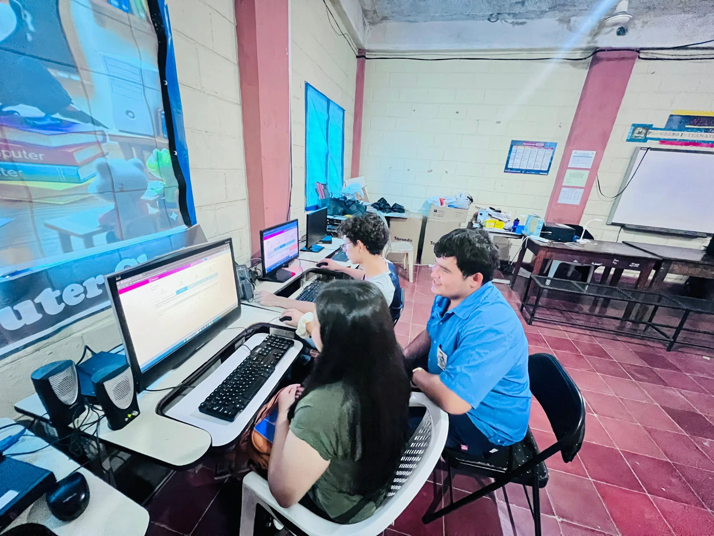 A classroom with students working on desktop computers. The room has a beige brick wall with pink accents, windows covered with blue curtains, and various items on the tables and behind them.