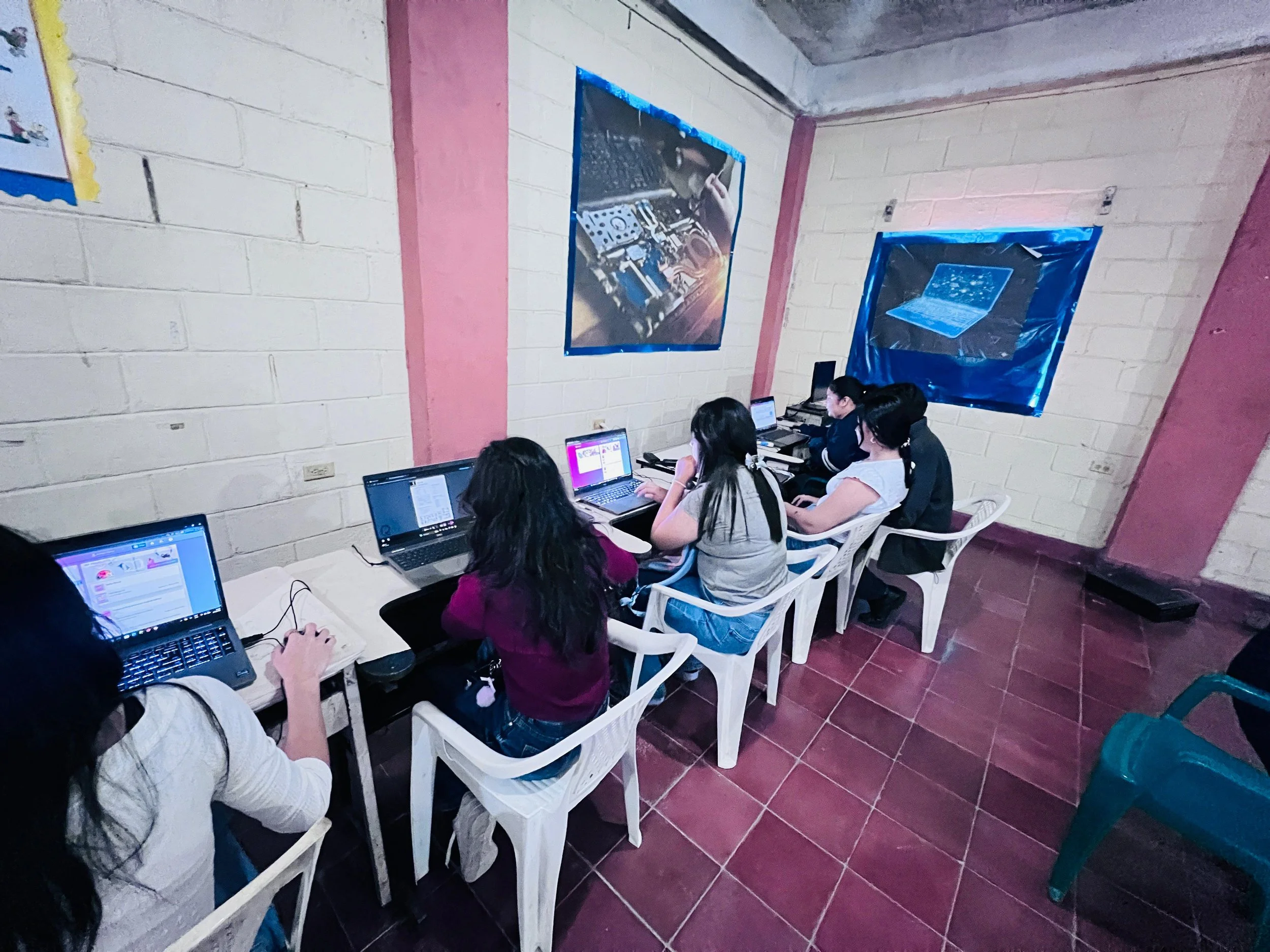 A classroom with students sitting at desks using laptops, with posters of technological equipment and a galaxy-themed image on the wall.