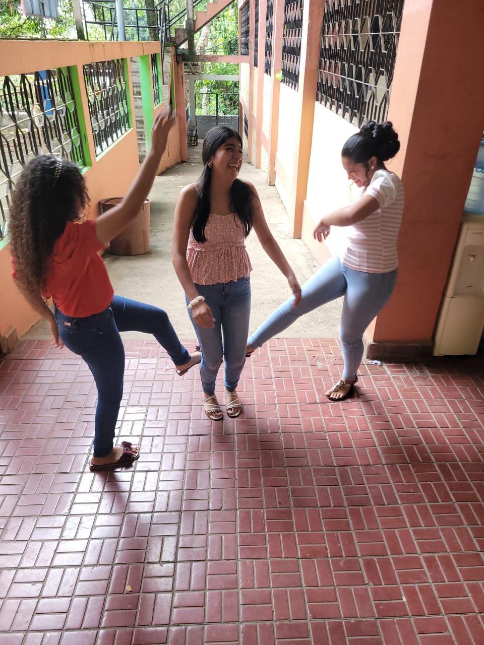 Three women laughing and playing together in a corridor, with one woman kicking her foot towards another's knee while the third woman stands between them, smiling.