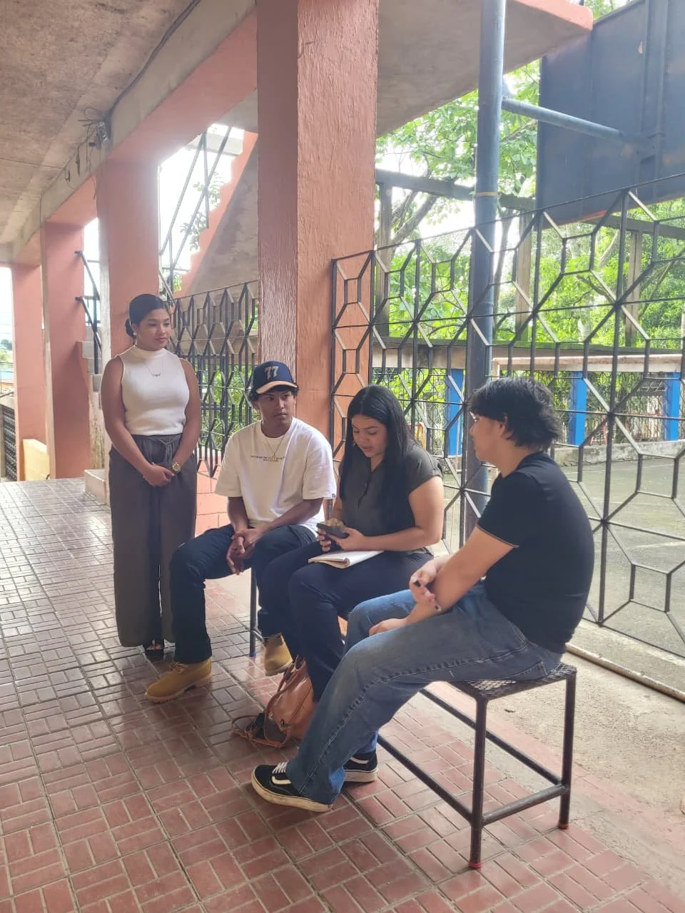 Four young people, two seated and two standing, in an outdoor area with a tiled floor, metal fencing, and visible green trees in the background.