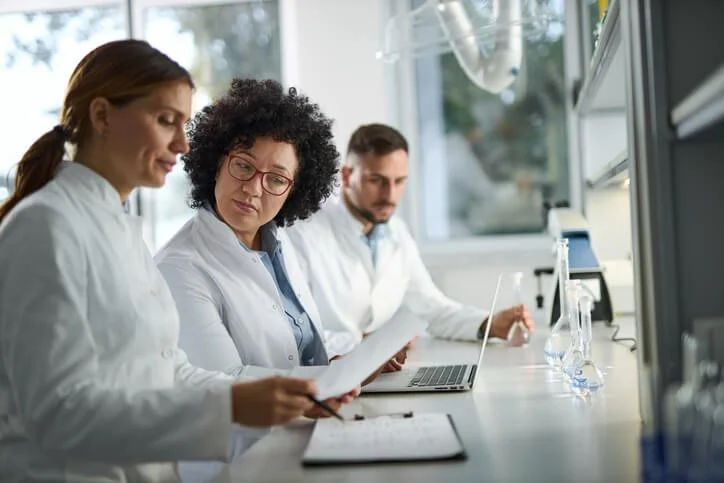 Three scientists in lab coats working in a laboratory, reviewing documents and working on a laptop, with scientific equipment on the counter.