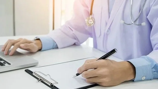 Healthcare professional wearing white coat and stethoscope writing on a clipboard with a pen, sitting at a desk with a laptop.