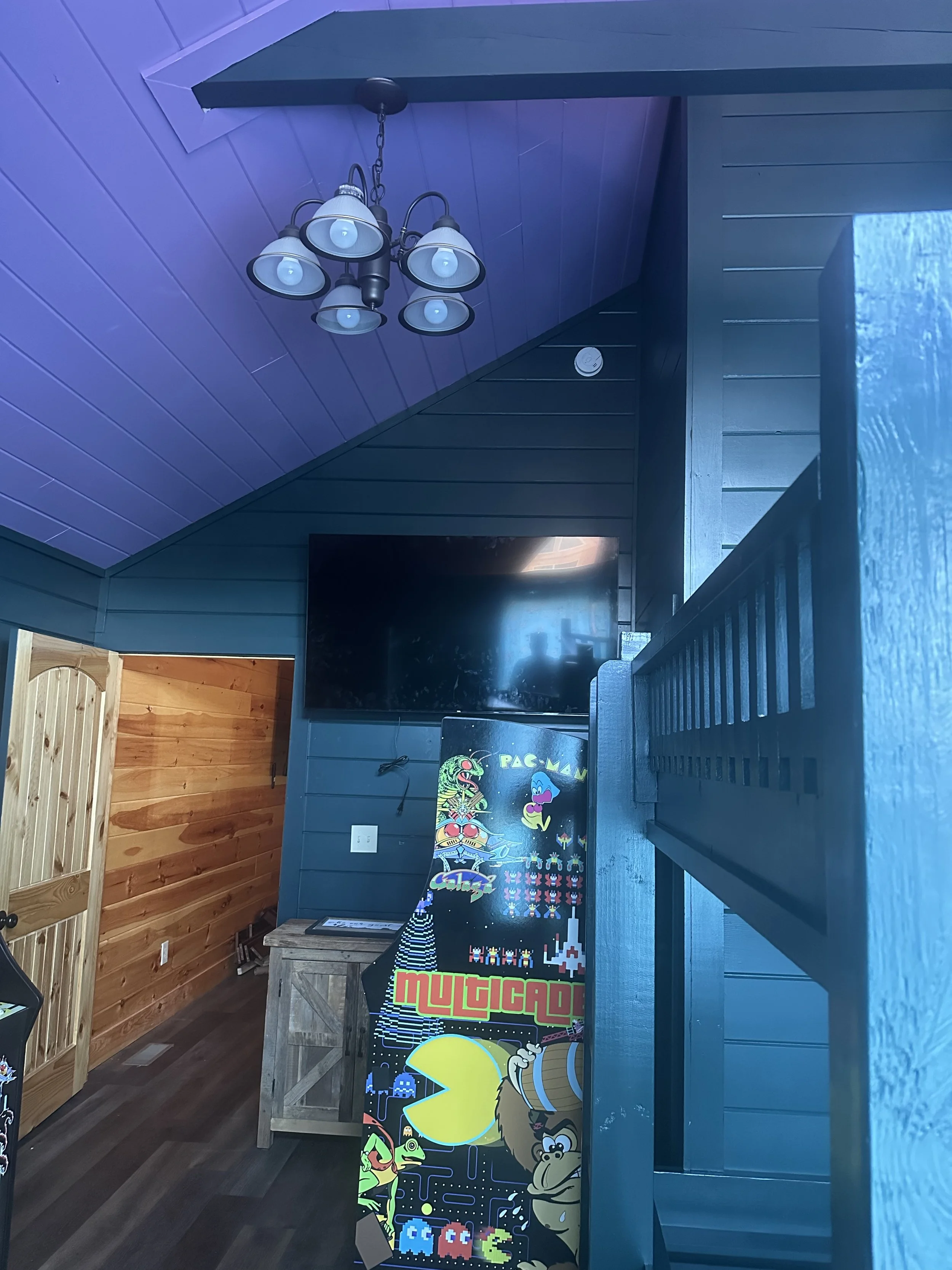 A corner of an indoor room with a ceiling lamp, a TV, a wooden table, a Pac-Man arcade game machine, and a blue wooden railing next to a window. The walls are paneled in dark and natural wood.