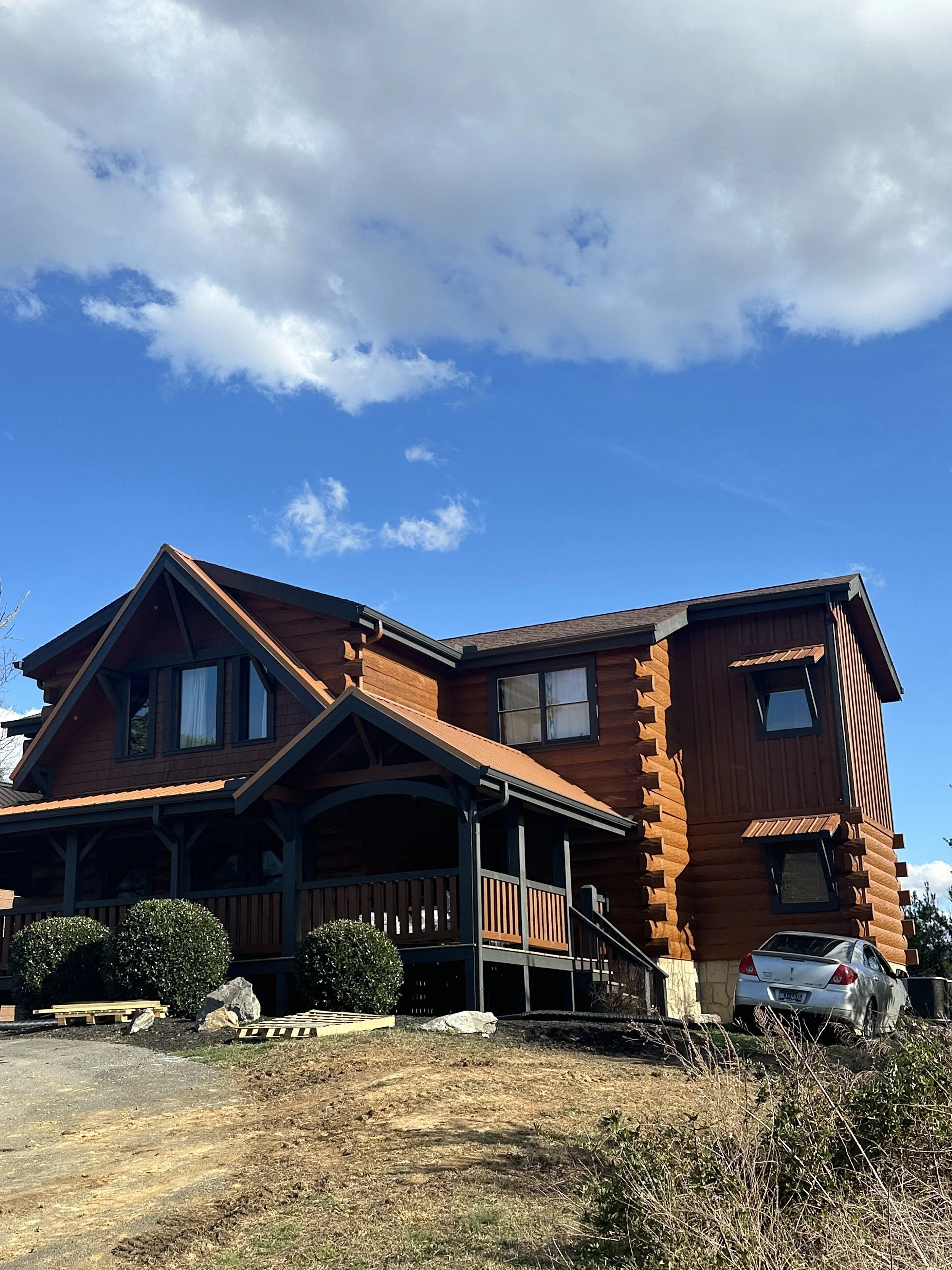 A two-story wooden house with a brown roof, multiple windows, and a porch, set against a bright blue sky with scattered white clouds. There are manicured bushes and a silver car parked nearby.