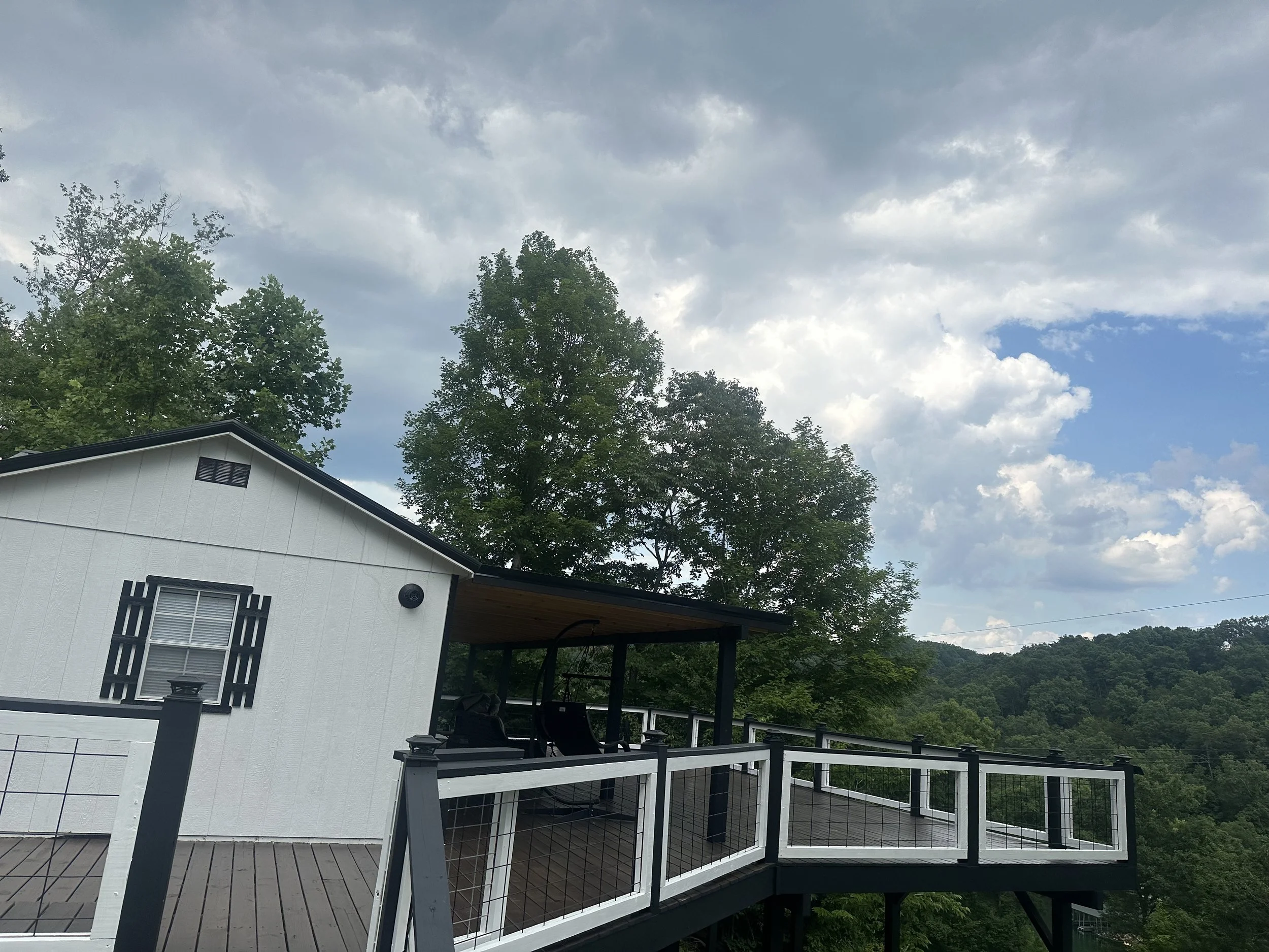 A house with a white exterior and black accents, surrounded by trees. It features a porch and a deck with black railings, overlooking a natural landscape with hills and cloudy sky.