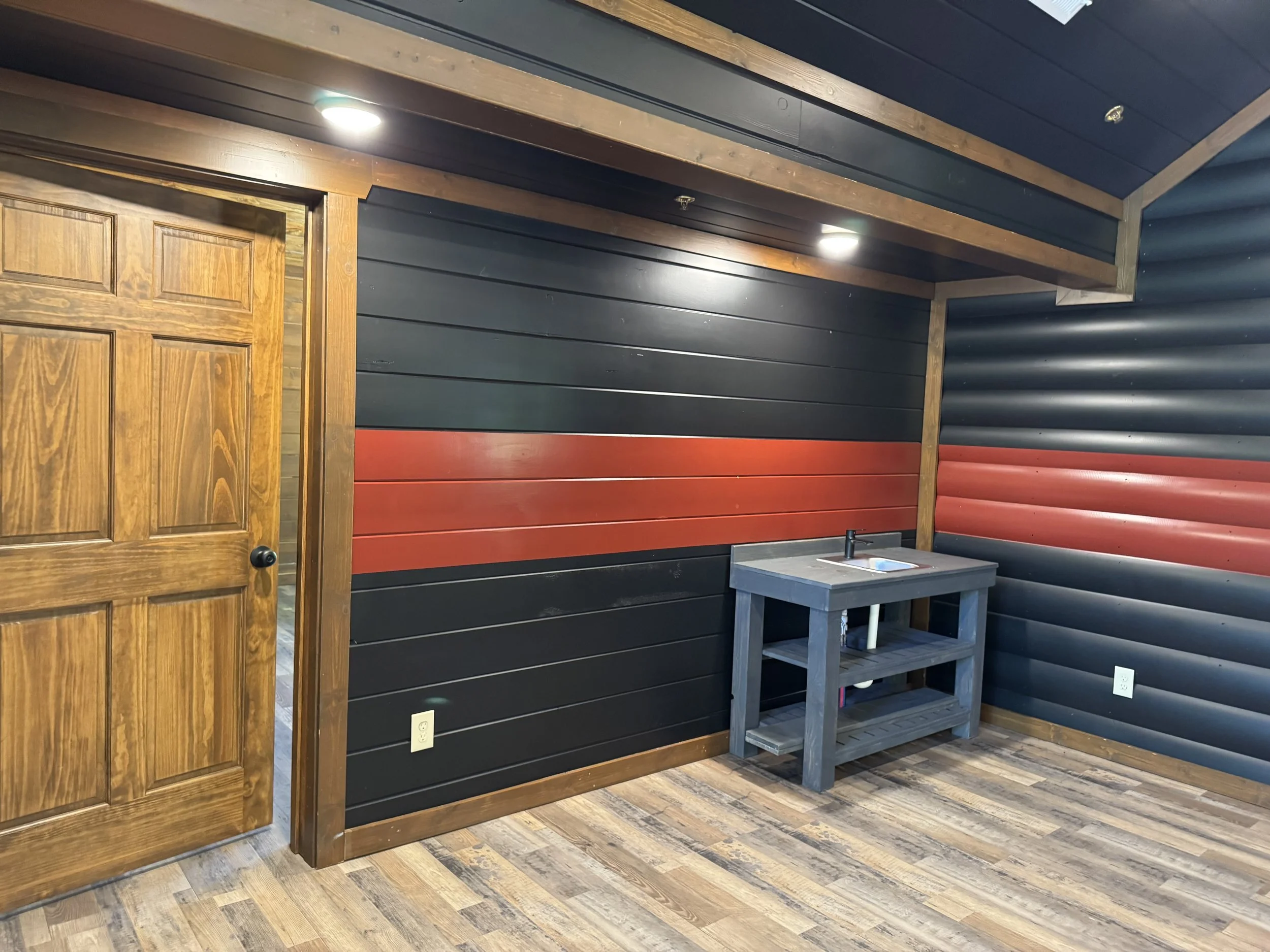 Interior of a room with dark horizontal wall panels, a wooden door on the left, a small gray table with a sink on the right, and wood flooring.