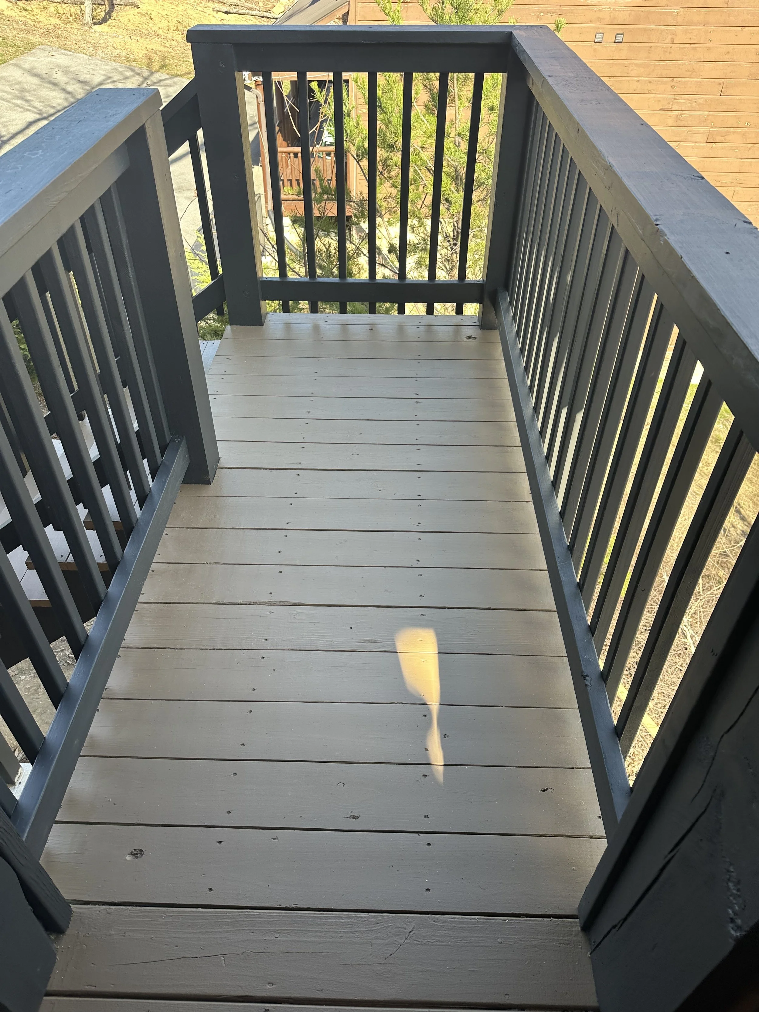 A small outdoor balcony with a wooden floor and black railings, overlooking a backyard with trees and a wooden fence.