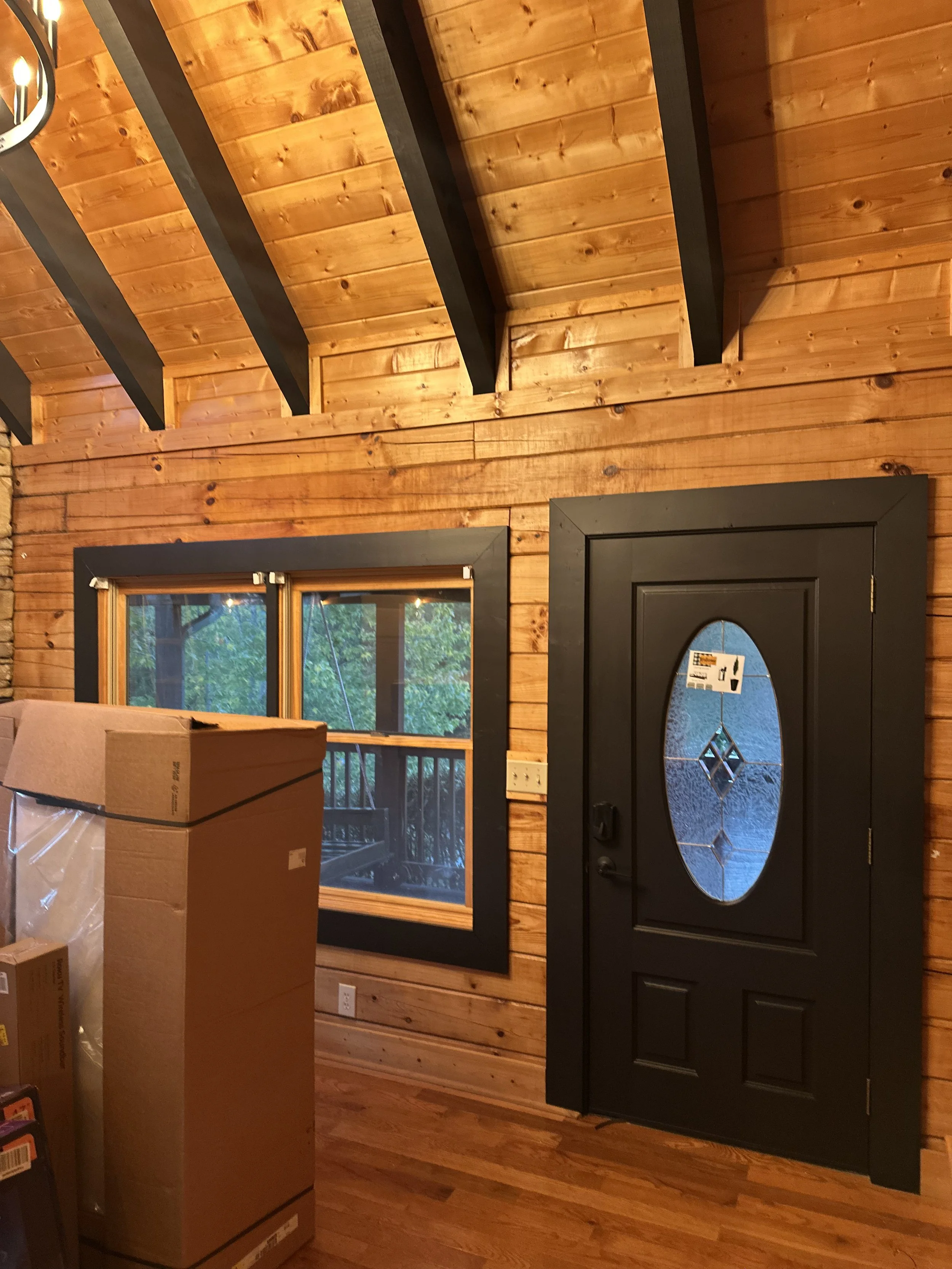 Interior of a wooden house with a black front door with a decorative glass oval, a window showing trees outside, and cardboard boxes inside.