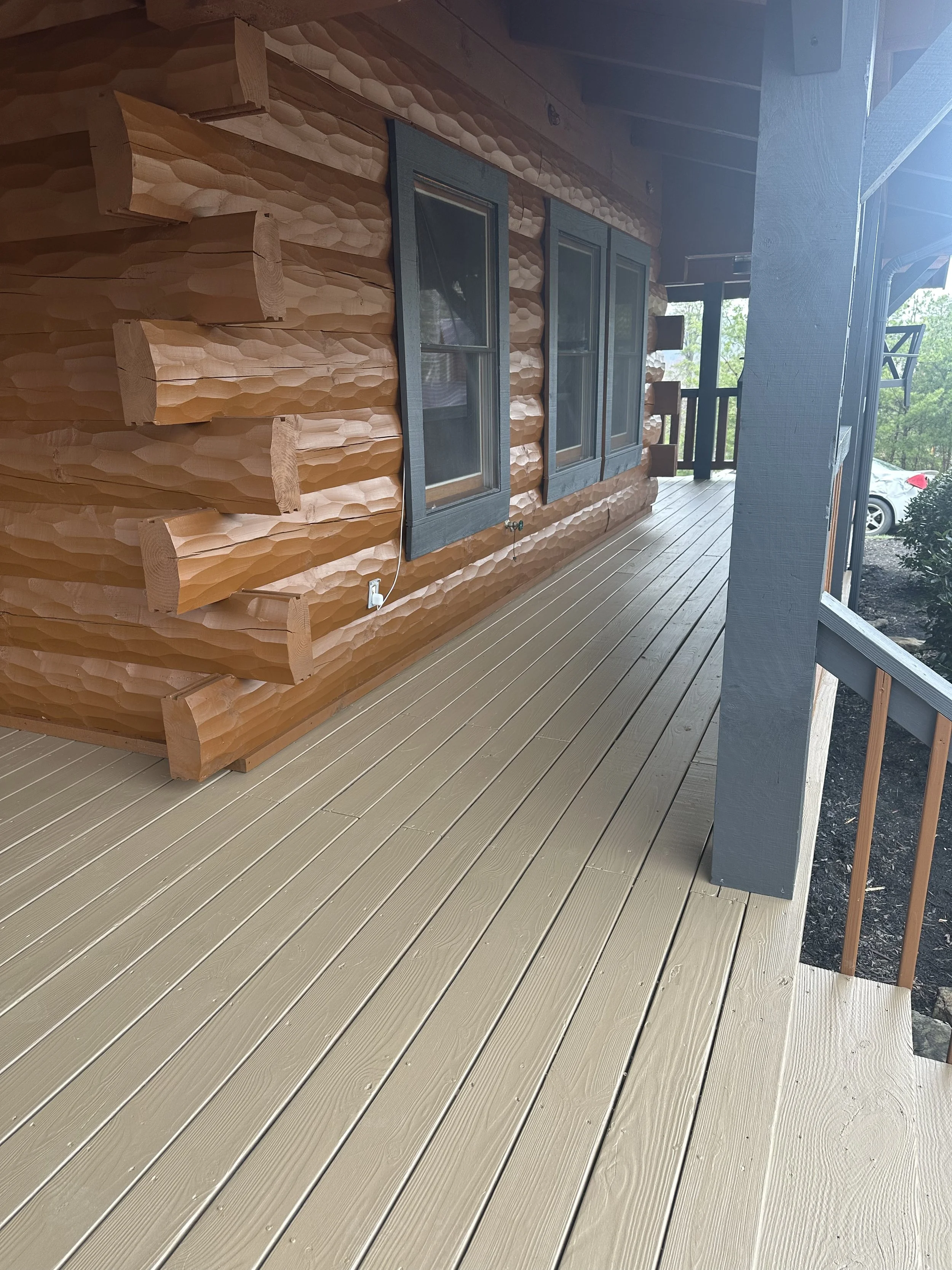 View of a porch with newly painted beige wooden floor boards and a log cabin wall with three blue window frames. There is a black railing and a car visible in the background.