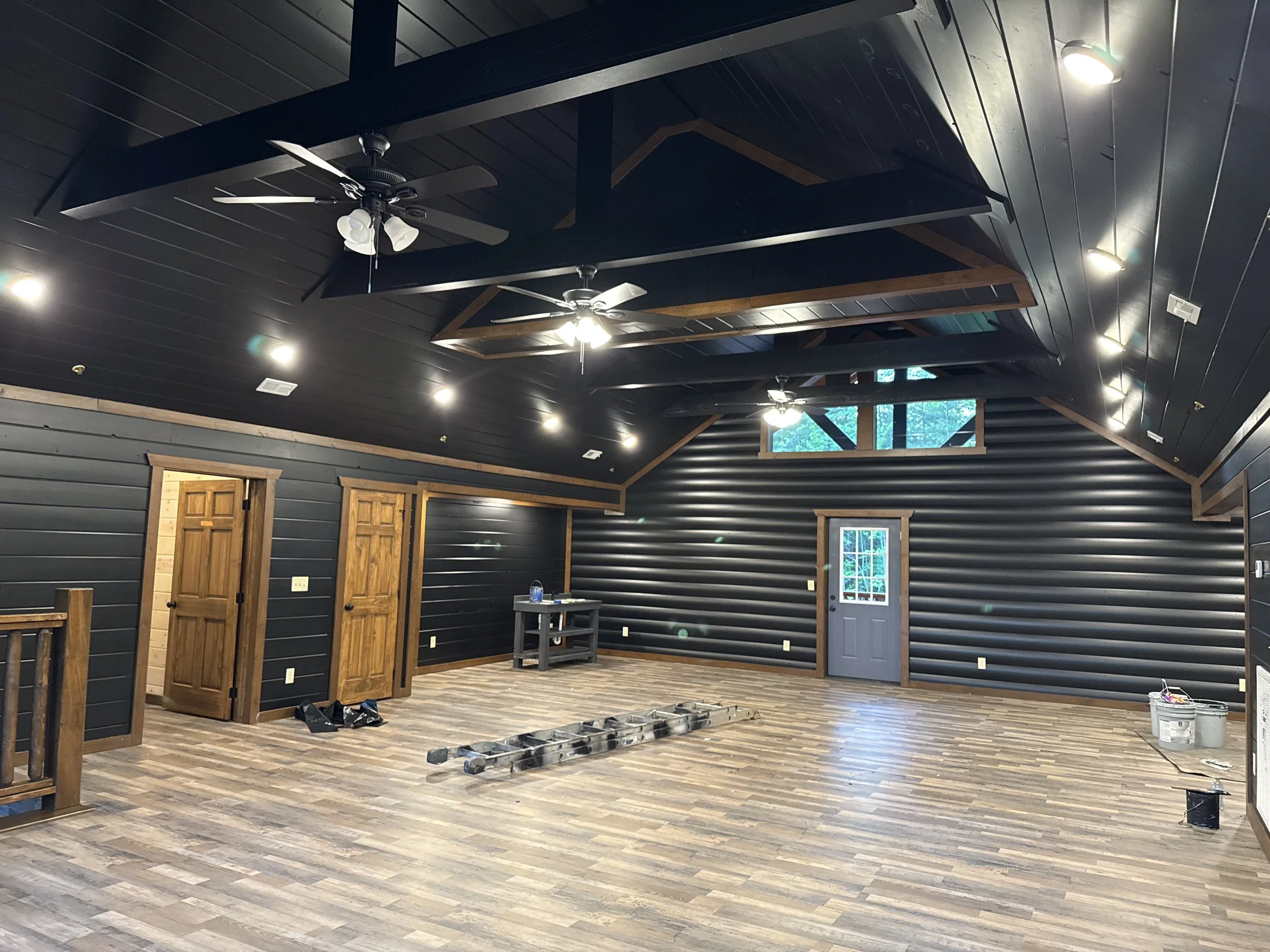 An interior room with black walls, wooden doors, and a high ceiling featuring ceiling fans and bright lights. The flooring is wooden, and some construction materials and tools are visible, indicating ongoing renovation.