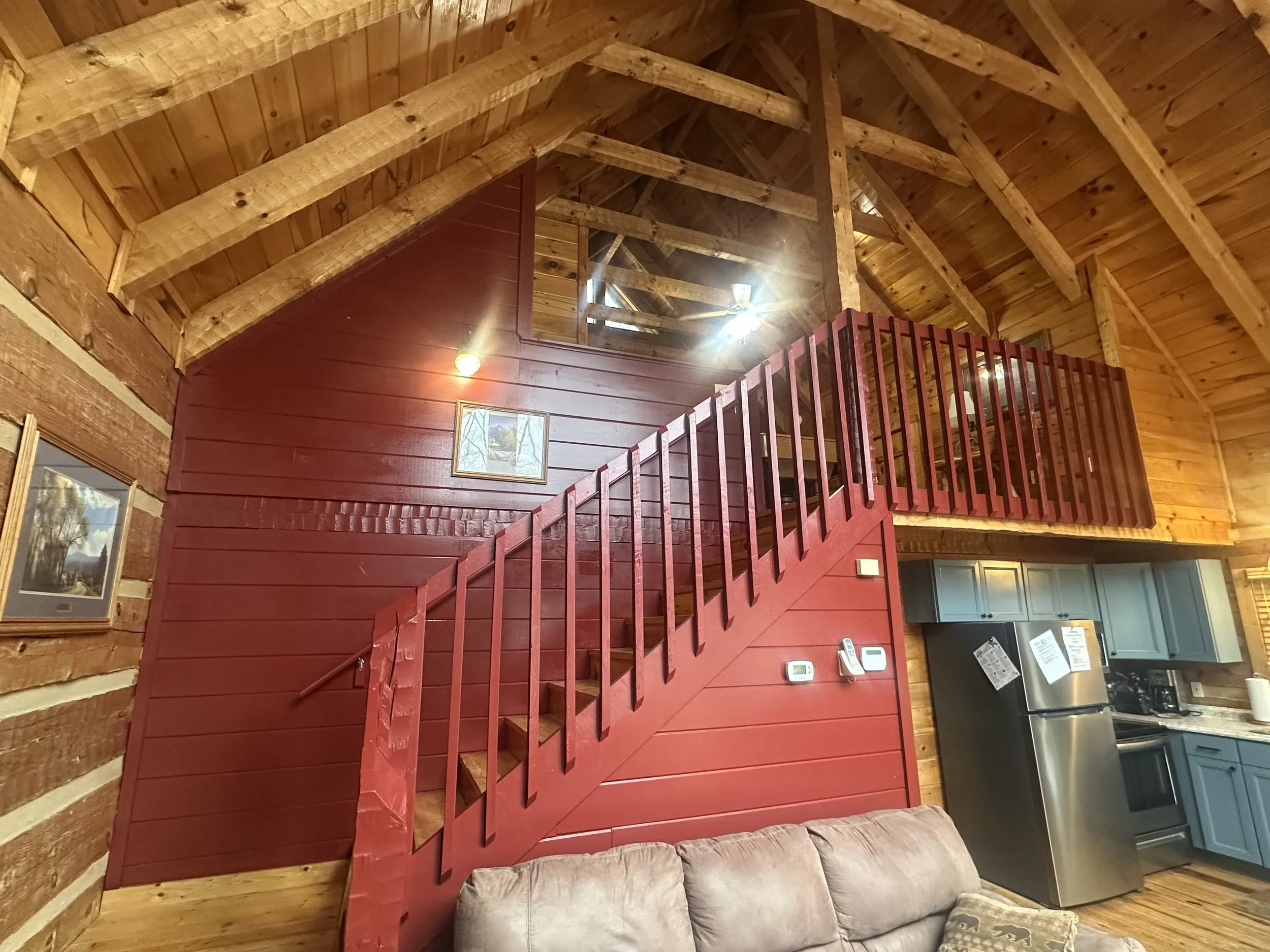 Interior of a log cabin with wooden walls, ceiling, and a red staircase leading to a loft area, decorated with home appliances and artwork.