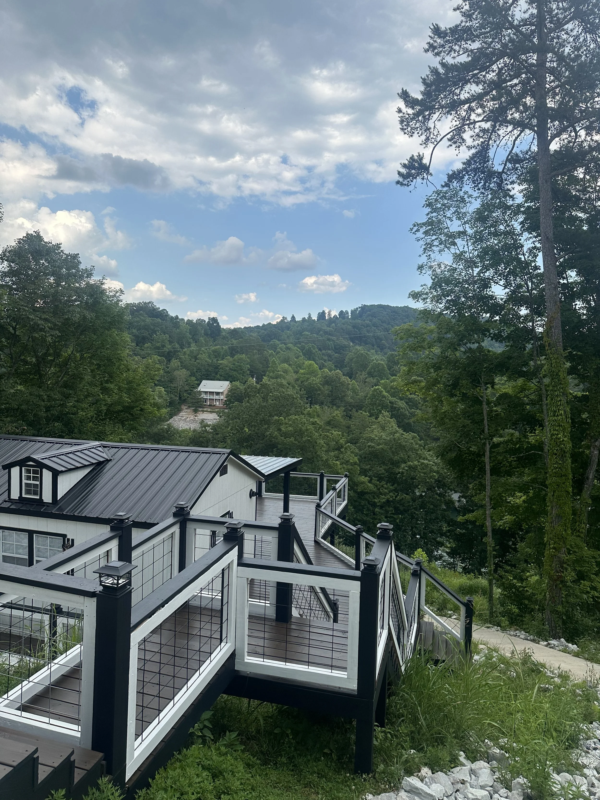 Balcony with black and white railing overlooking a lush green hillside with trees and a cloudy sky.