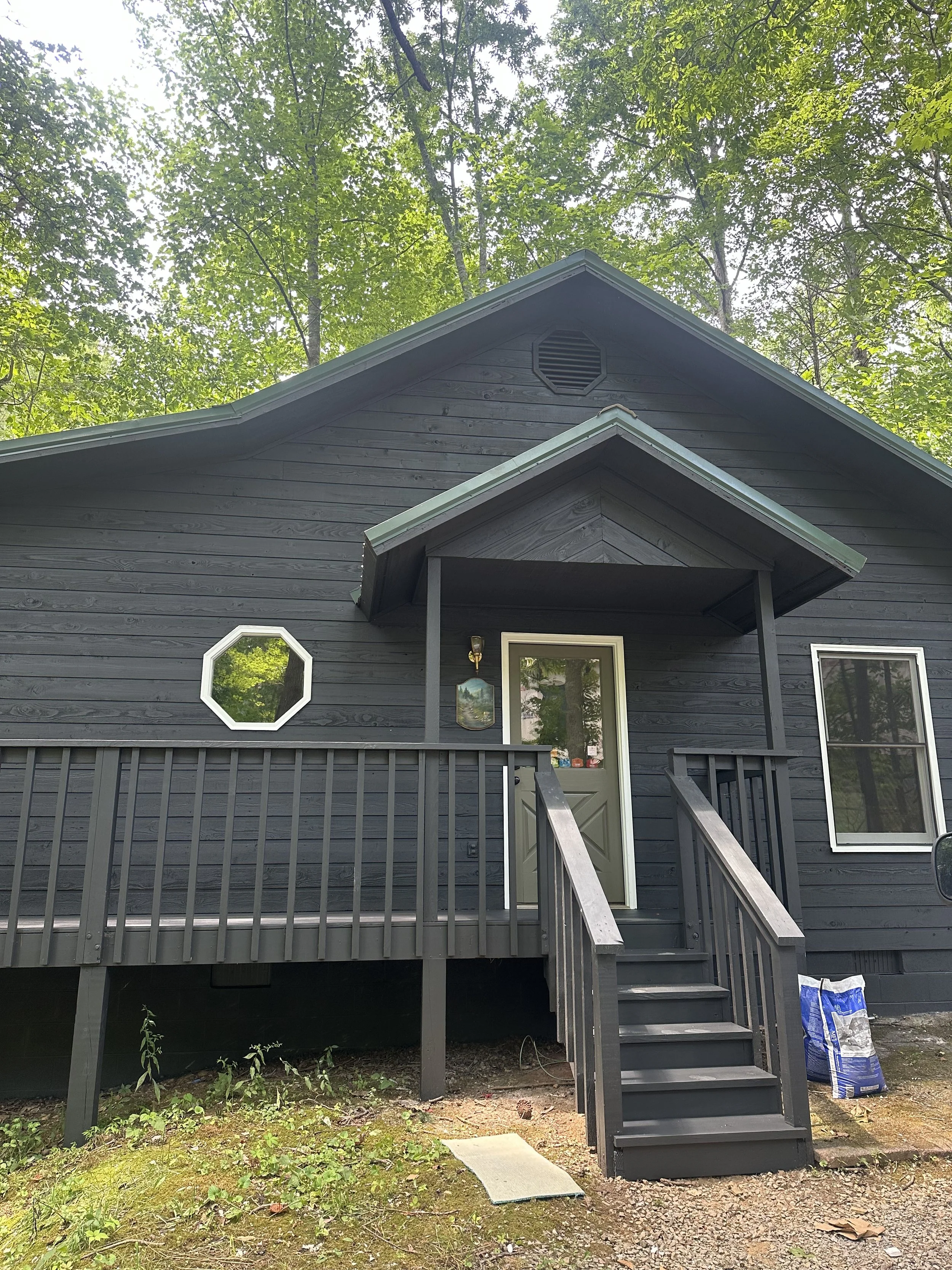 Front view of a dark gray house with a small porch and staircase, surrounded by green trees in the background and gardening supplies on the ground.