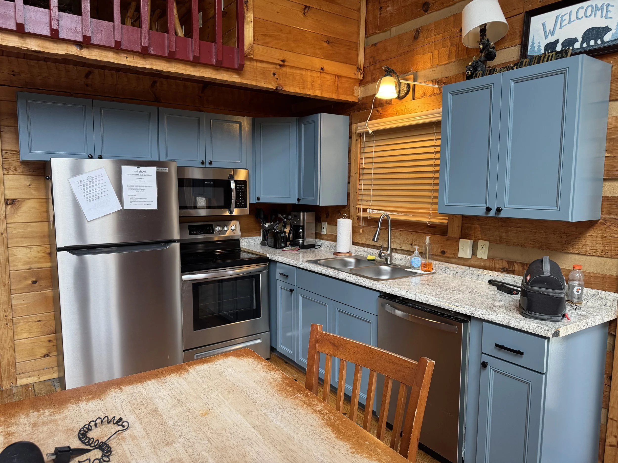 A rustic kitchen with wooden walls and ceiling, featuring blue cabinets, a stainless steel refrigerator, a microwave, and a granite countertop. A window with closed blinds, various small appliances, and decorative bear figurines are also visible.