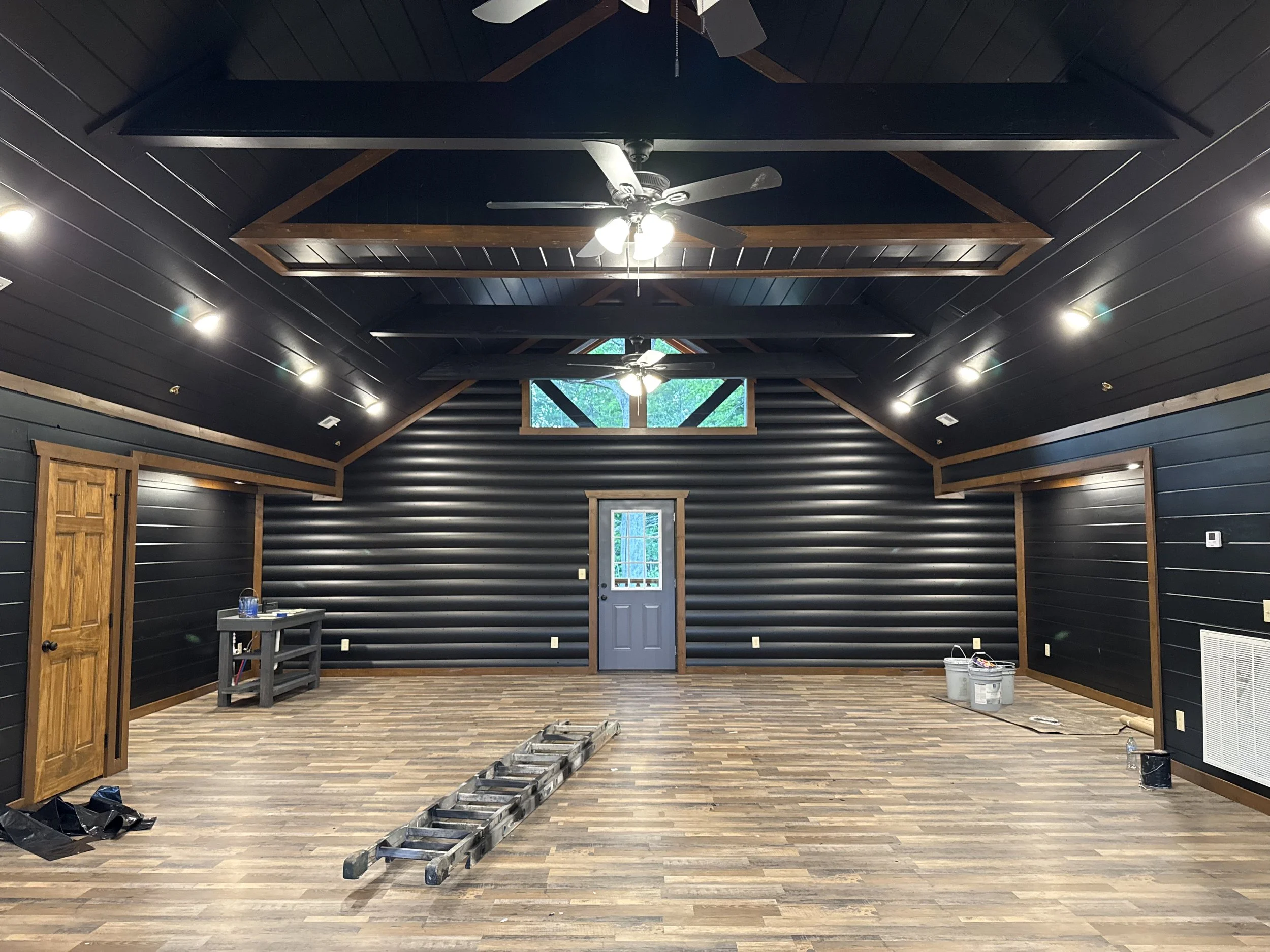 Empty room with black paneled walls and ceiling, wooden beams, and a ceiling fan with lights. There are windows near the ceiling and a door at the far wall. Construction materials and tools are scattered on the floor.