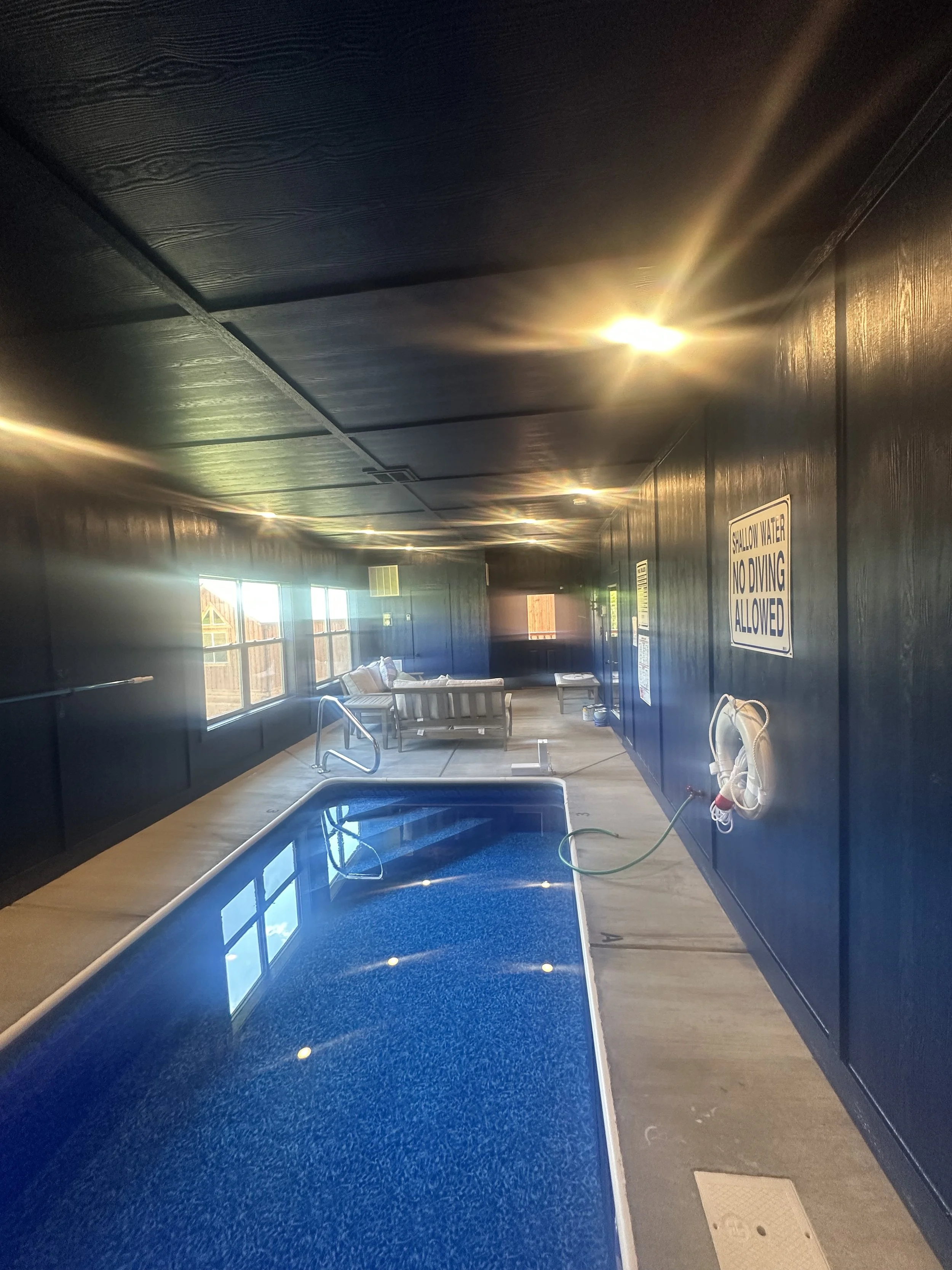 Indoor swimming pool with blue water and a black ceiling. Lounge chairs and a life preserver are on the side, with a sign that reads 'Shallow Water No Diving Allowed' on the wall.