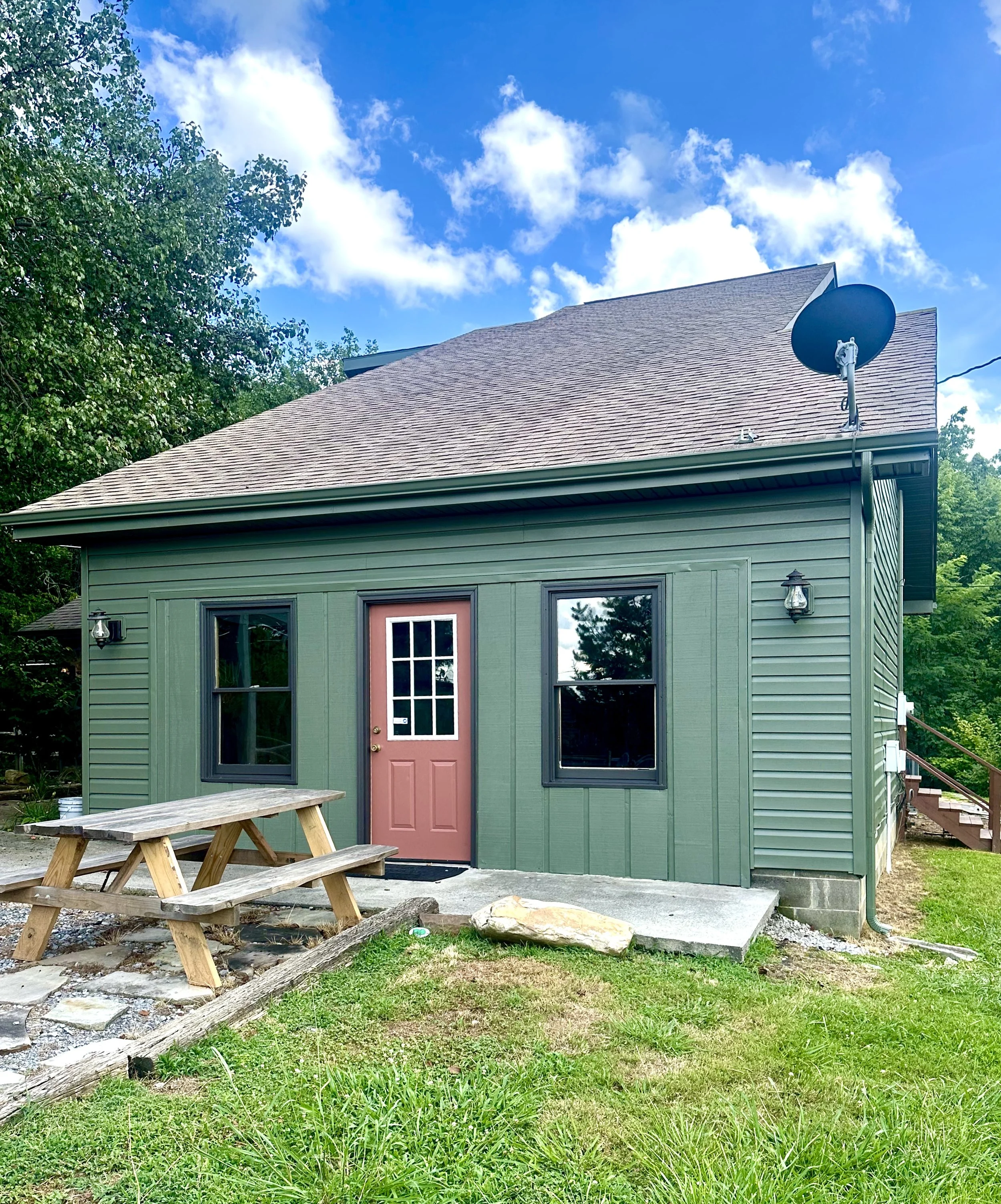 A house with green exterior siding and a pink door, surrounded by green grass and trees under a partly cloudy sky. There is a satellite dish mounted on the roof, a lantern mounted on the wall near the door, and a picnic table in the yard.