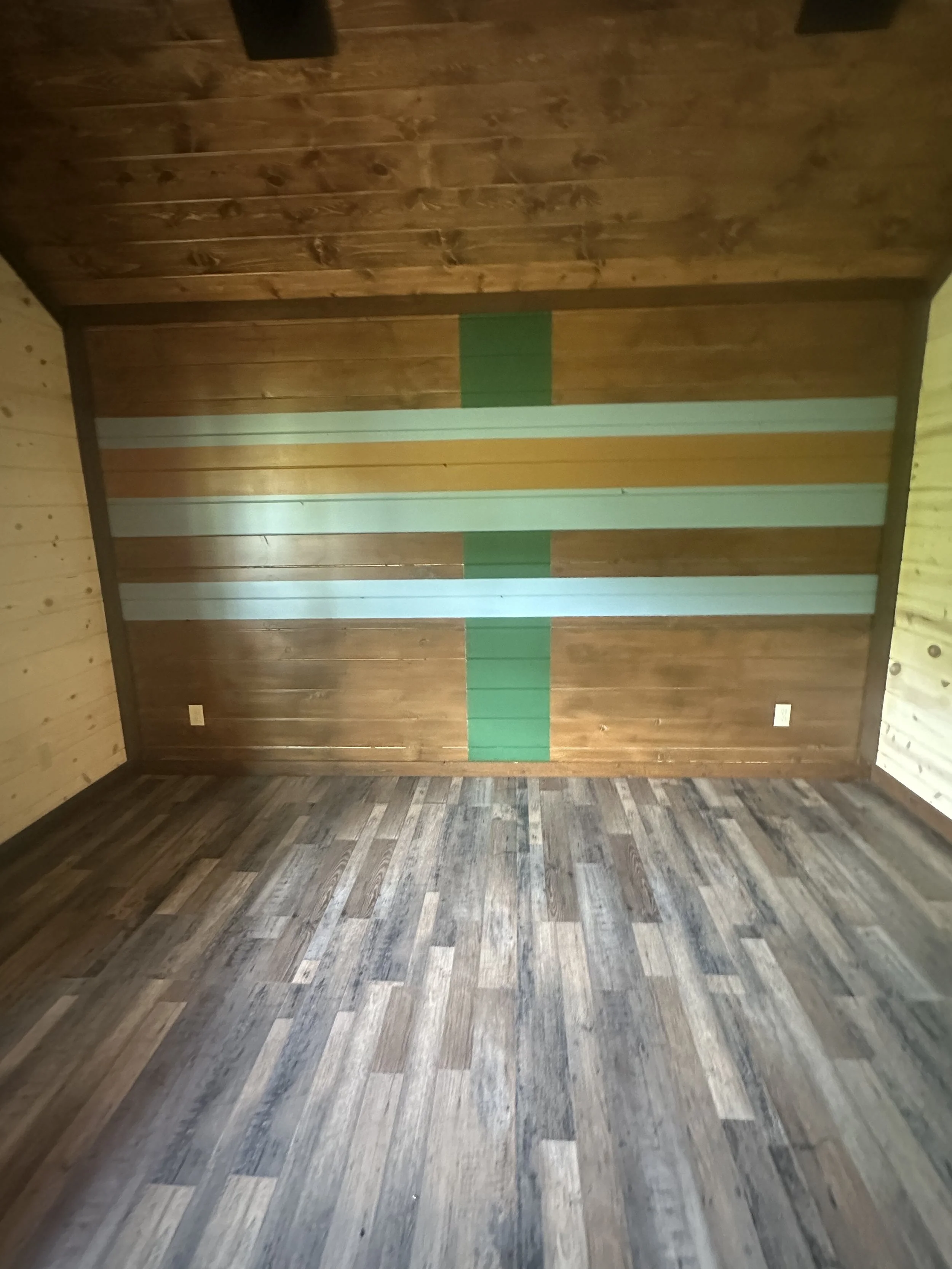 Photo of an interior wooden room with a wooden floor and a wall decorated with horizontal colorful wooden planks, including shades of brown, white, yellow, and green.