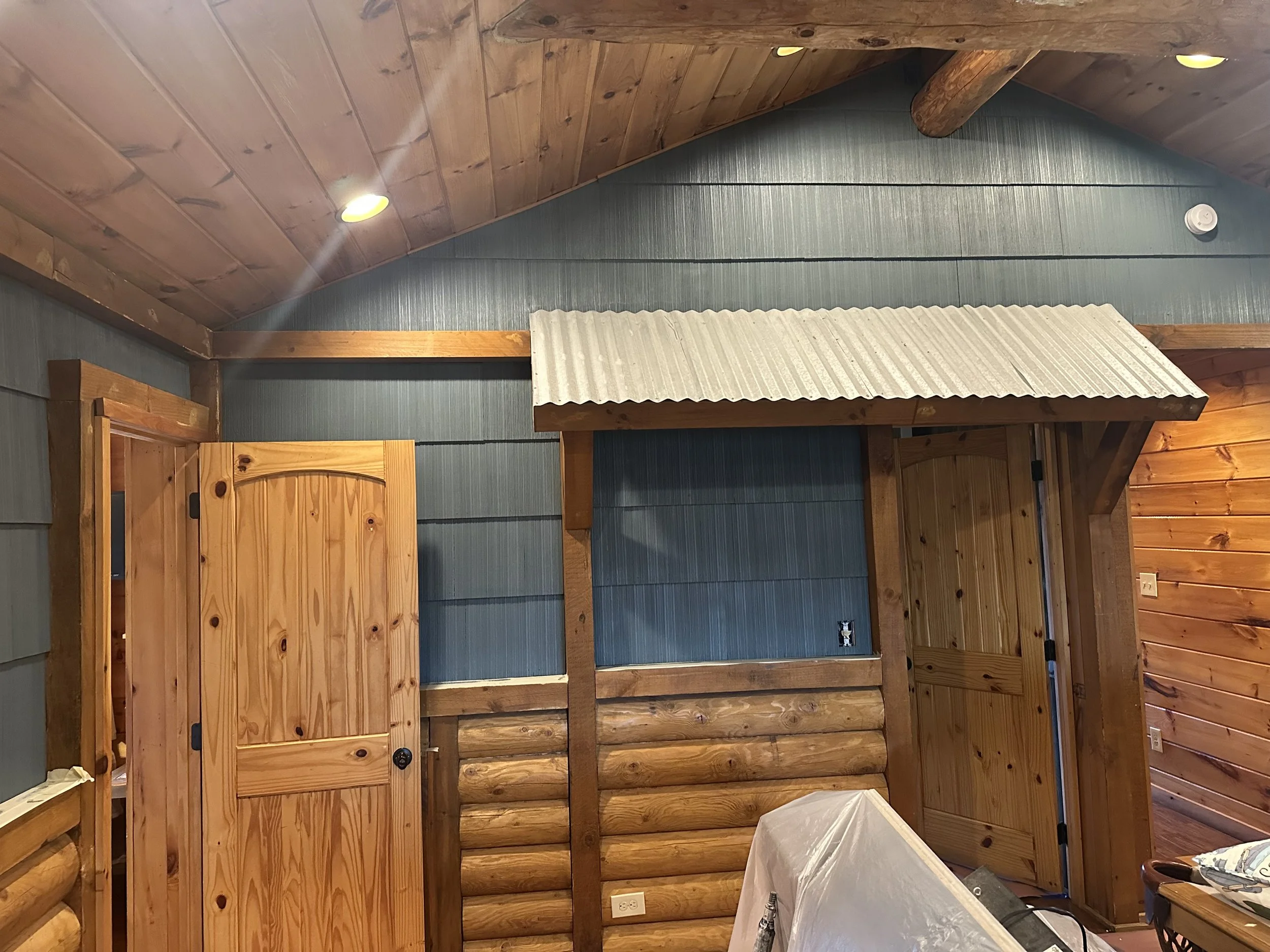 Interior of a wooden cabin or room with a vaulted wooden ceiling, wooden beams, and paneled walls, some of which are painted blue. There are two recessed lights in the ceiling, a smoke detector on the wall, a closed wooden door, and a covered object in the foreground.