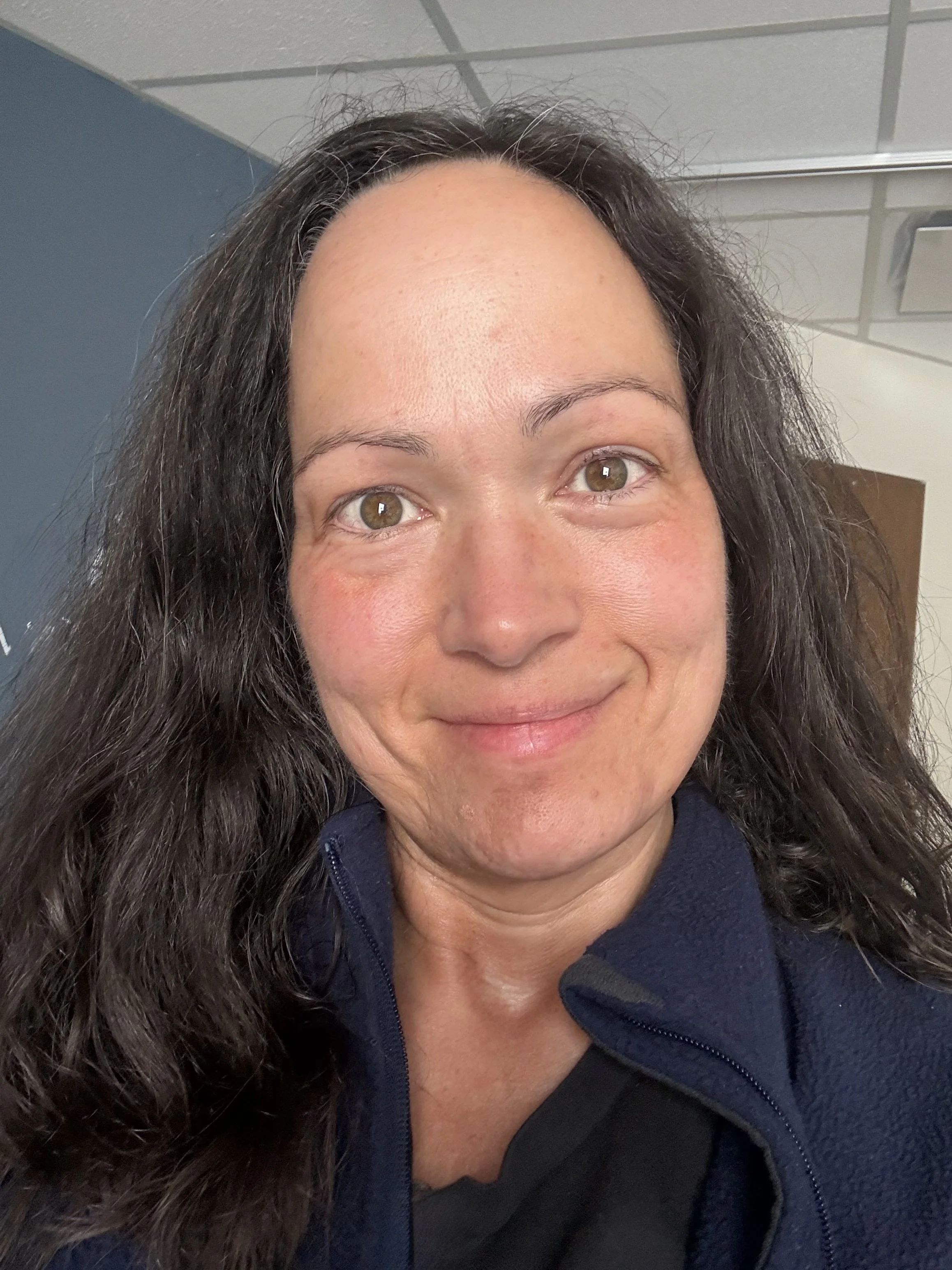 A woman with long, dark hair and hazel eyes smiling in a close-up photo, wearing a dark zippered jacket.