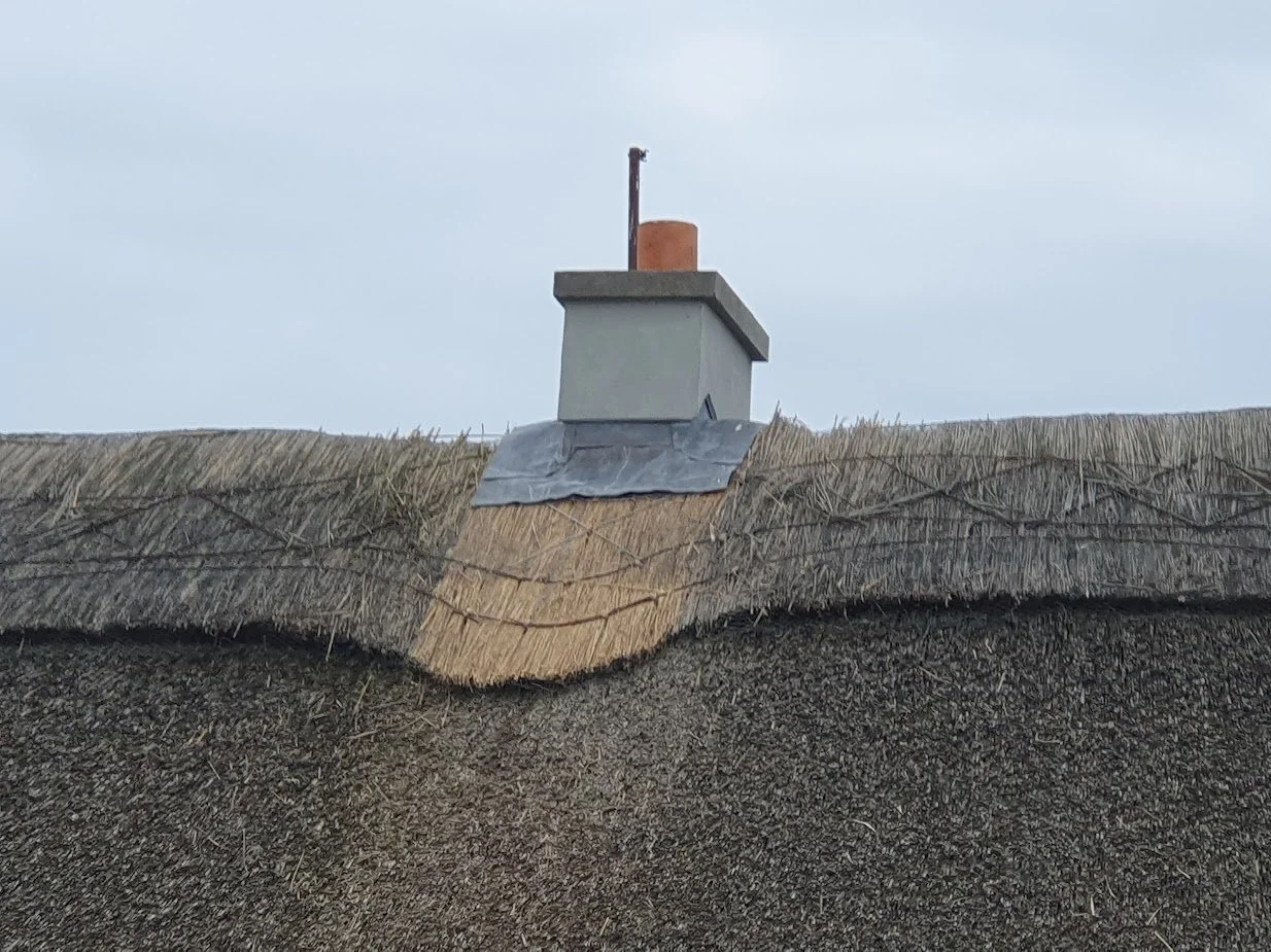 Close-up of a roof with a chimney, thatched roofing, and some metal flashing around the chimney.