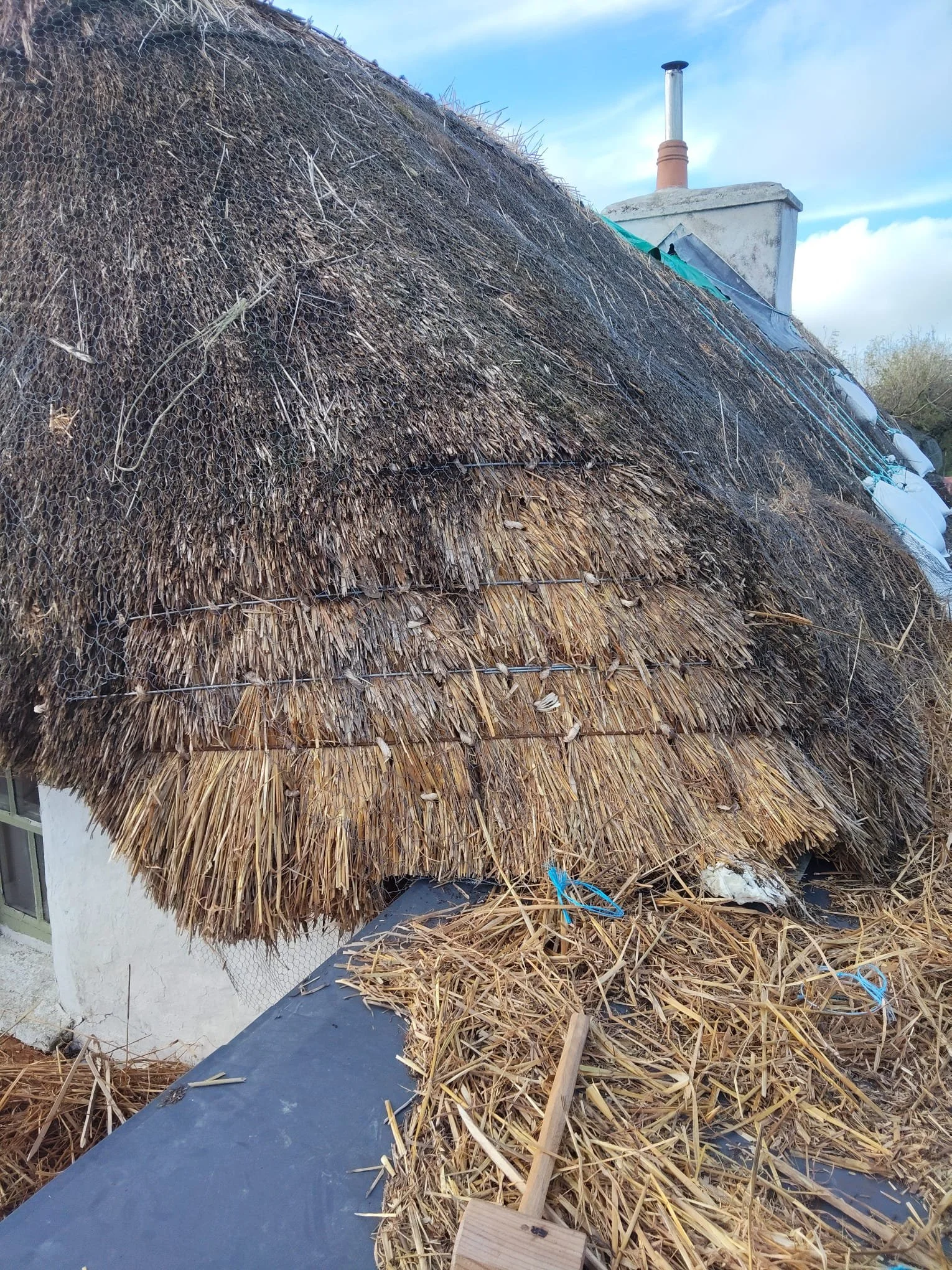Thatched roof with straw and twigs, part of a white building, with a chimney on top, and some construction debris and straw scattered on the roof.