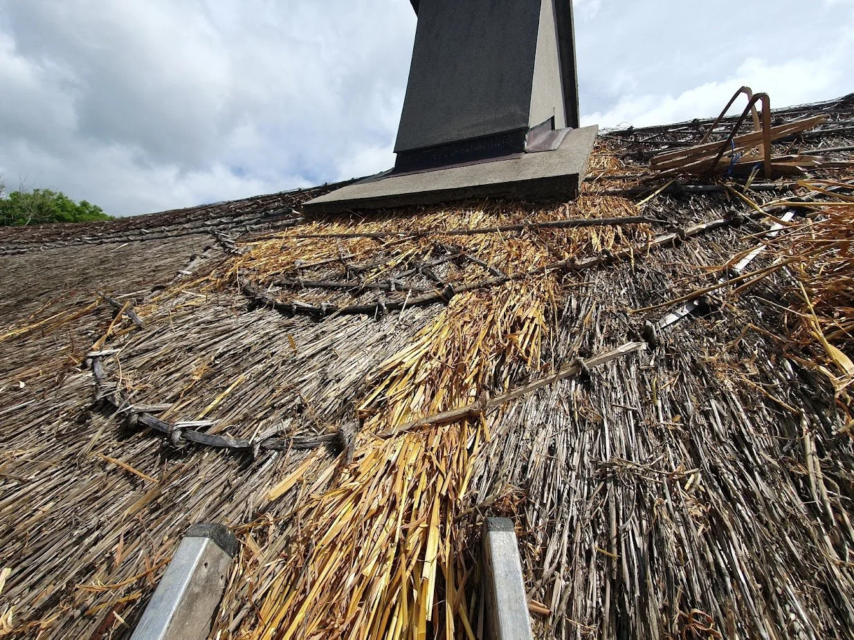 Close-up of a thatched roof with a chimney, showing damaged and burnt thatching material with a ladder in the foreground.
