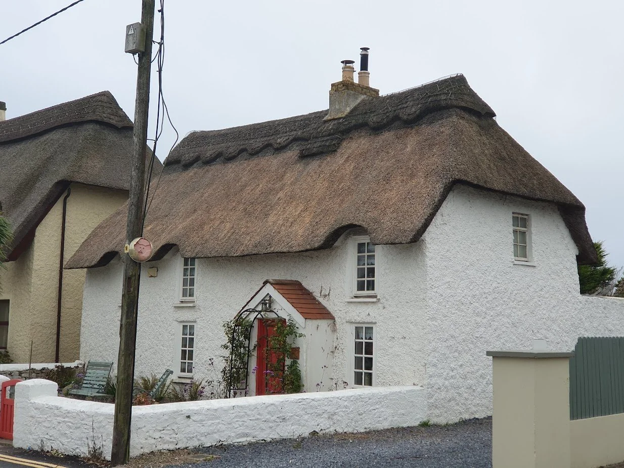 A white cottage with a thatched roof and small windows, with a red front door and a garden area.