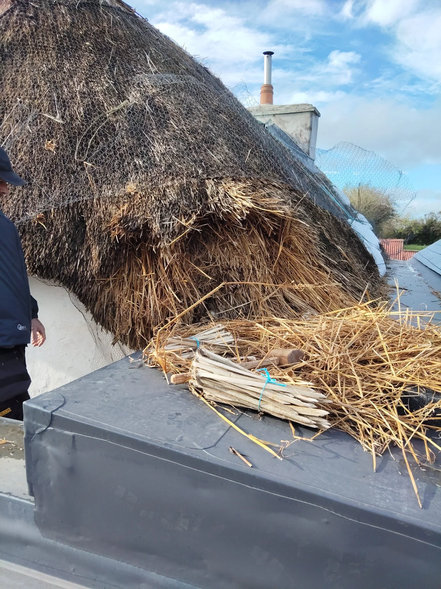 A large heap of thatched roofing material and bamboo sticks on top of a black vehicle, with some mesh netting and a chimney visible in the background under a cloudy sky.