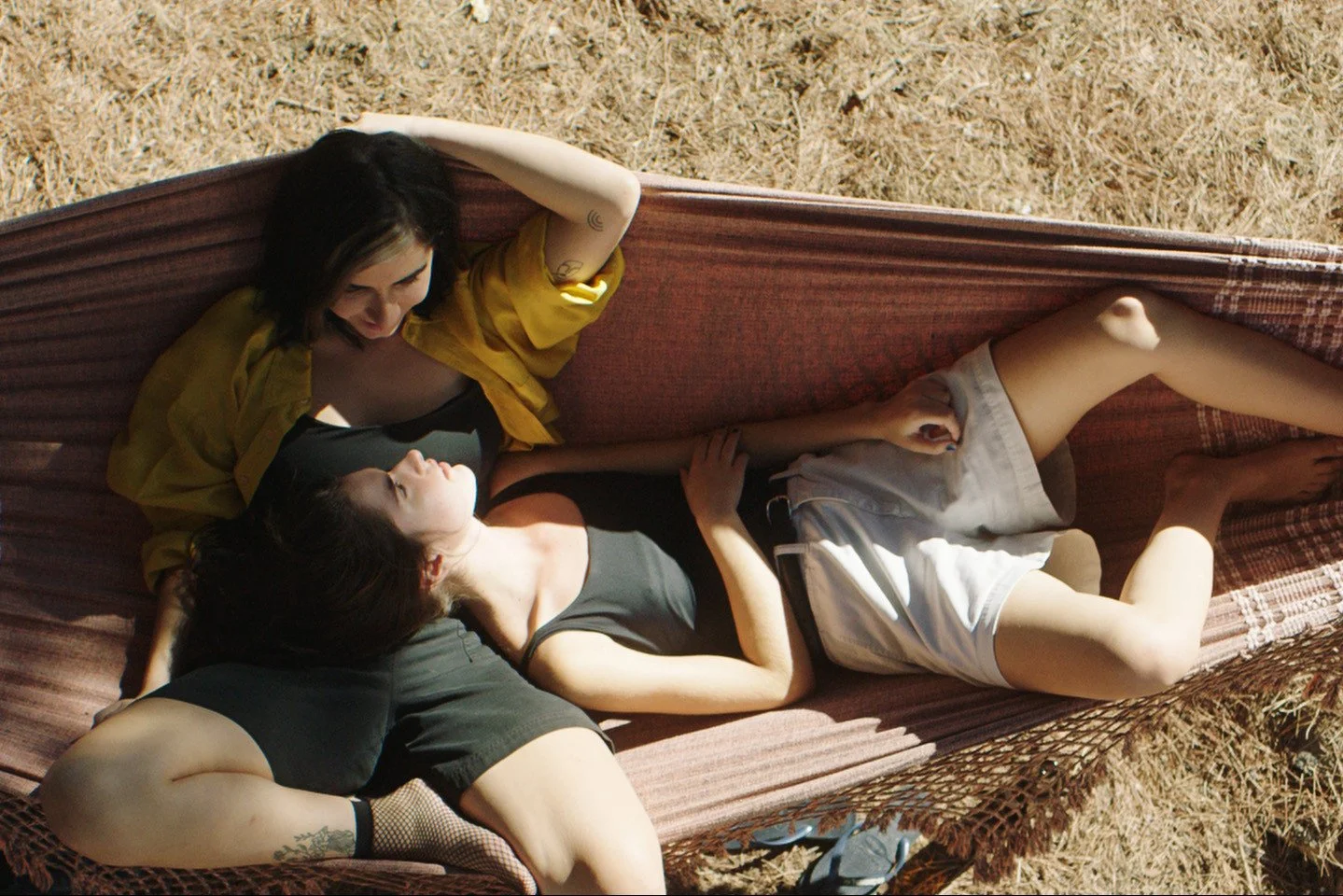 Three women lying on a lounge chair outdoors, two looking into each other's eyes, under bright sunlight.