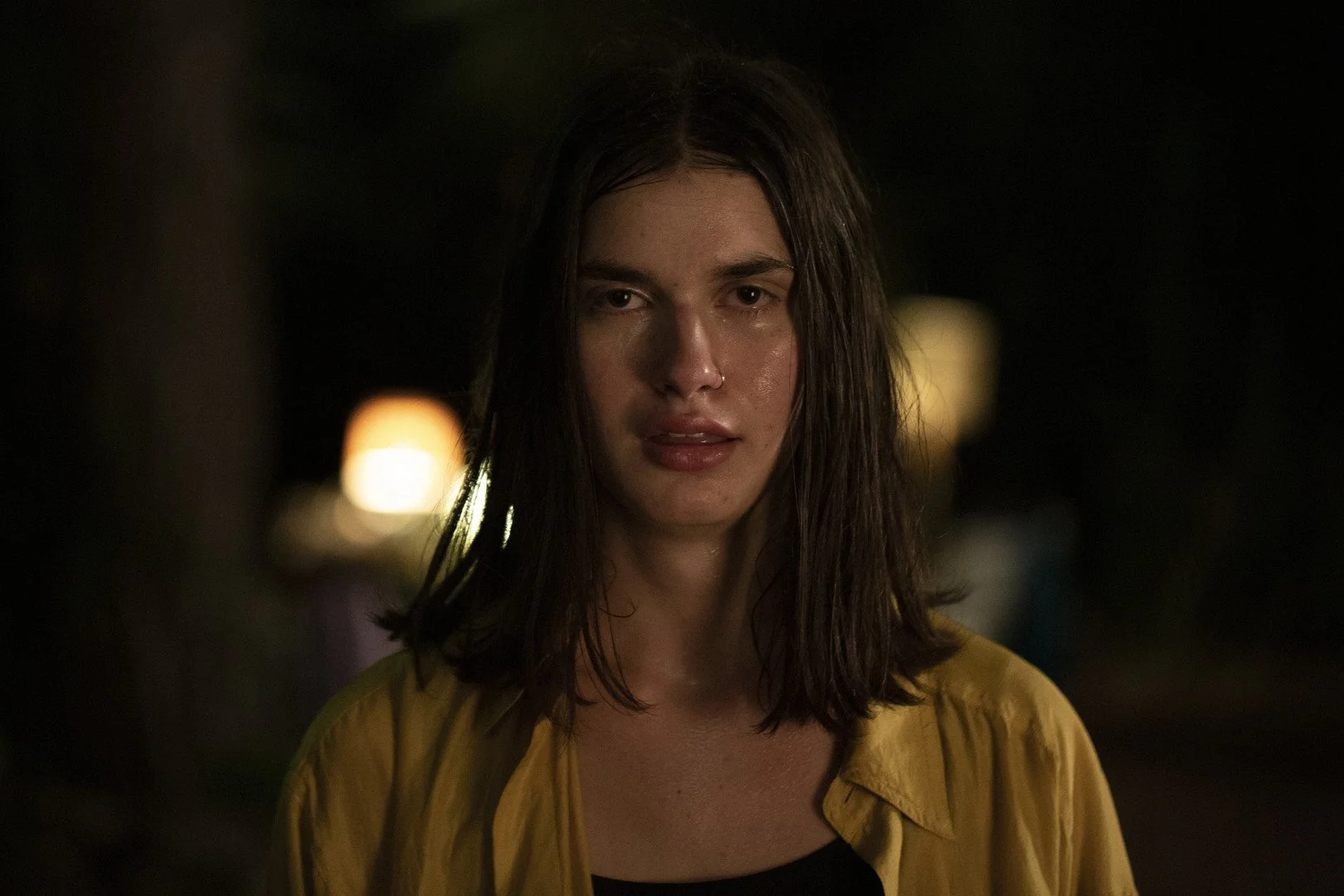A woman with wet hair and a yellow jacket looks directly at the camera at night.